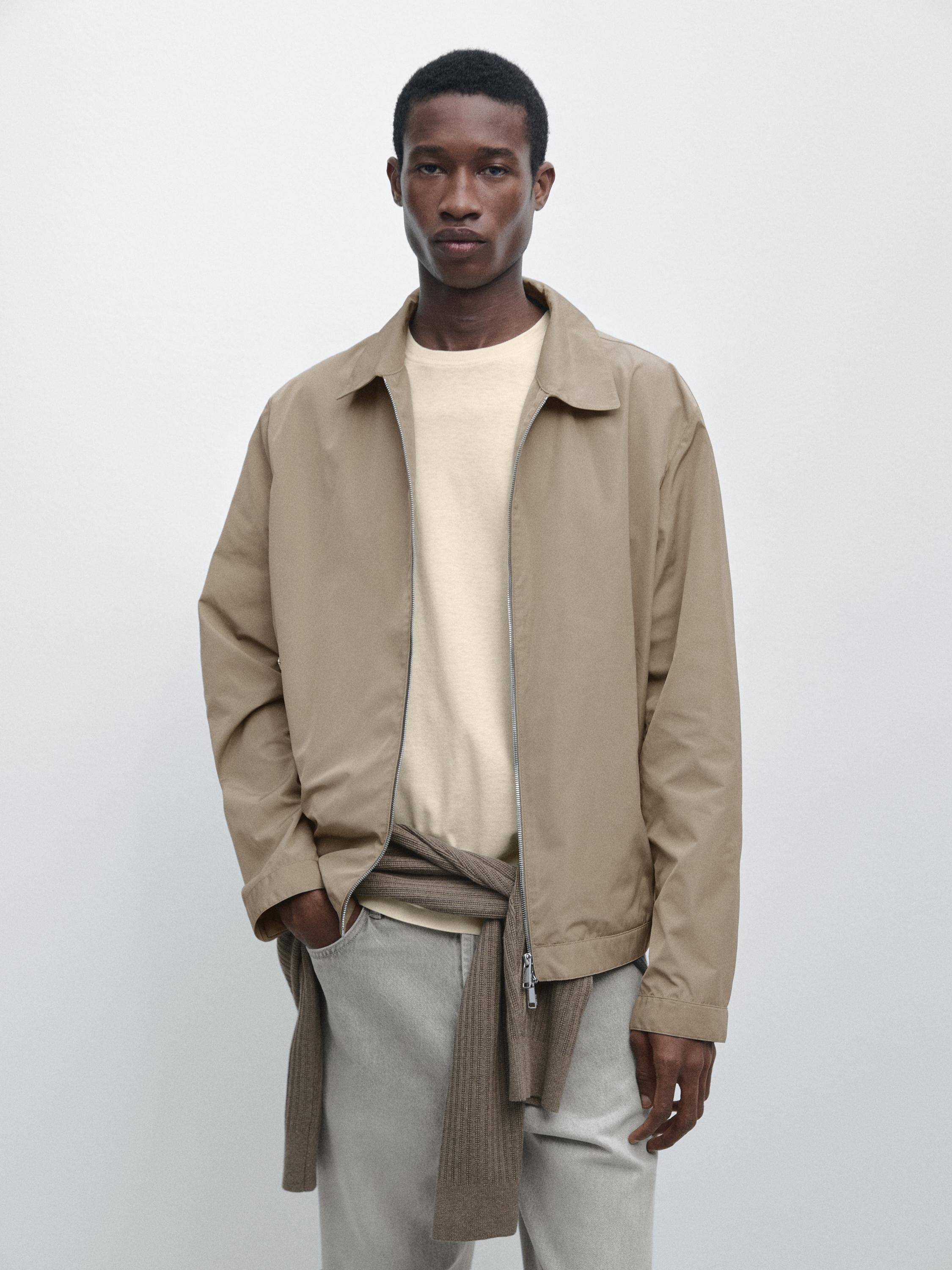 Kurze Nerzjacke mit Reißverschluss, beiges T-Shirt und graue Hose. Brauner Pullover um die Taille gebunden.