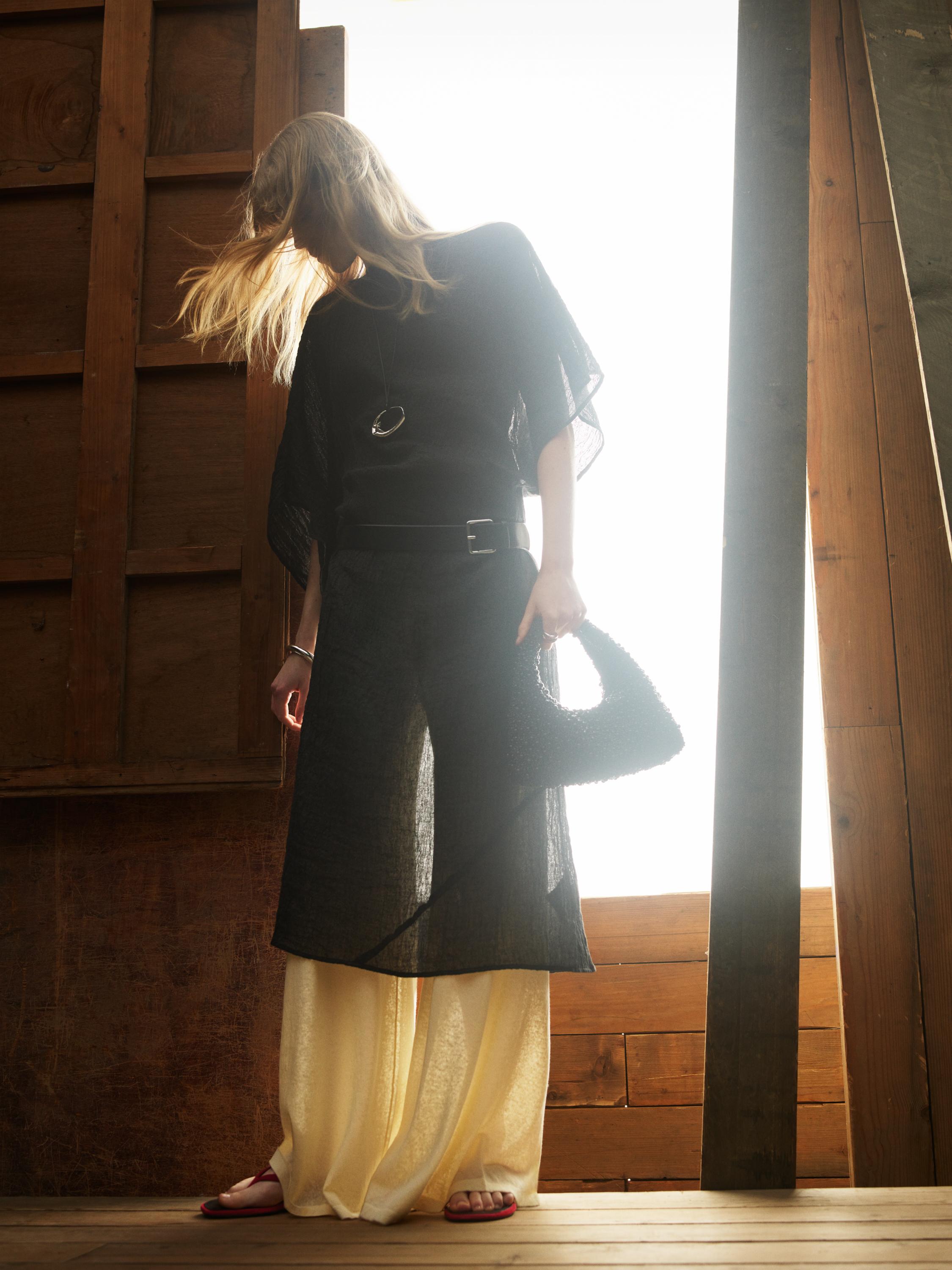 Σύνολο από ημιδιάφανη μαύρη μπλούζα και απαλό παντελόνι βανίλια. Φοράει κόκκινα σανδάλια και μαύρη τσάντα.