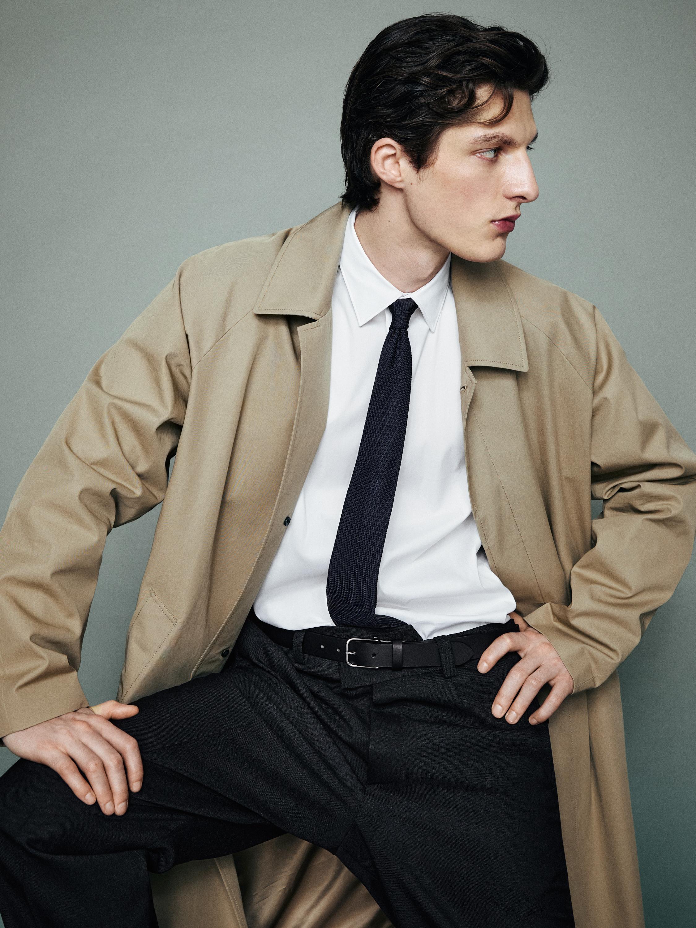 Ensemble avec gabardine beige, chemise blanche, cravate noire et pantalon foncé avec ceinture noire.