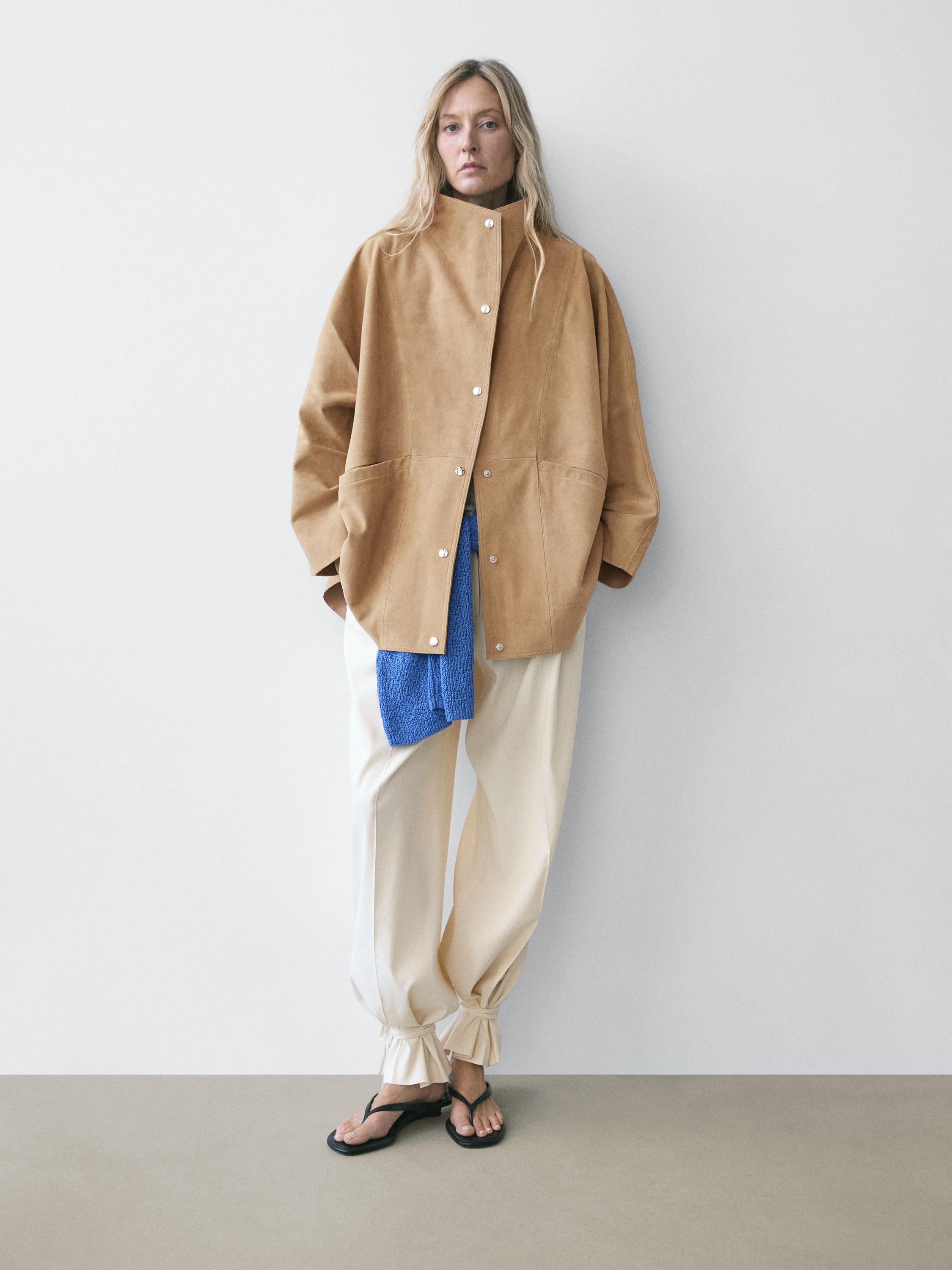 Zandkleurige jas set met knopen en voorzakken, beige broek met strikken bij de enkels en blauwe sjaal.