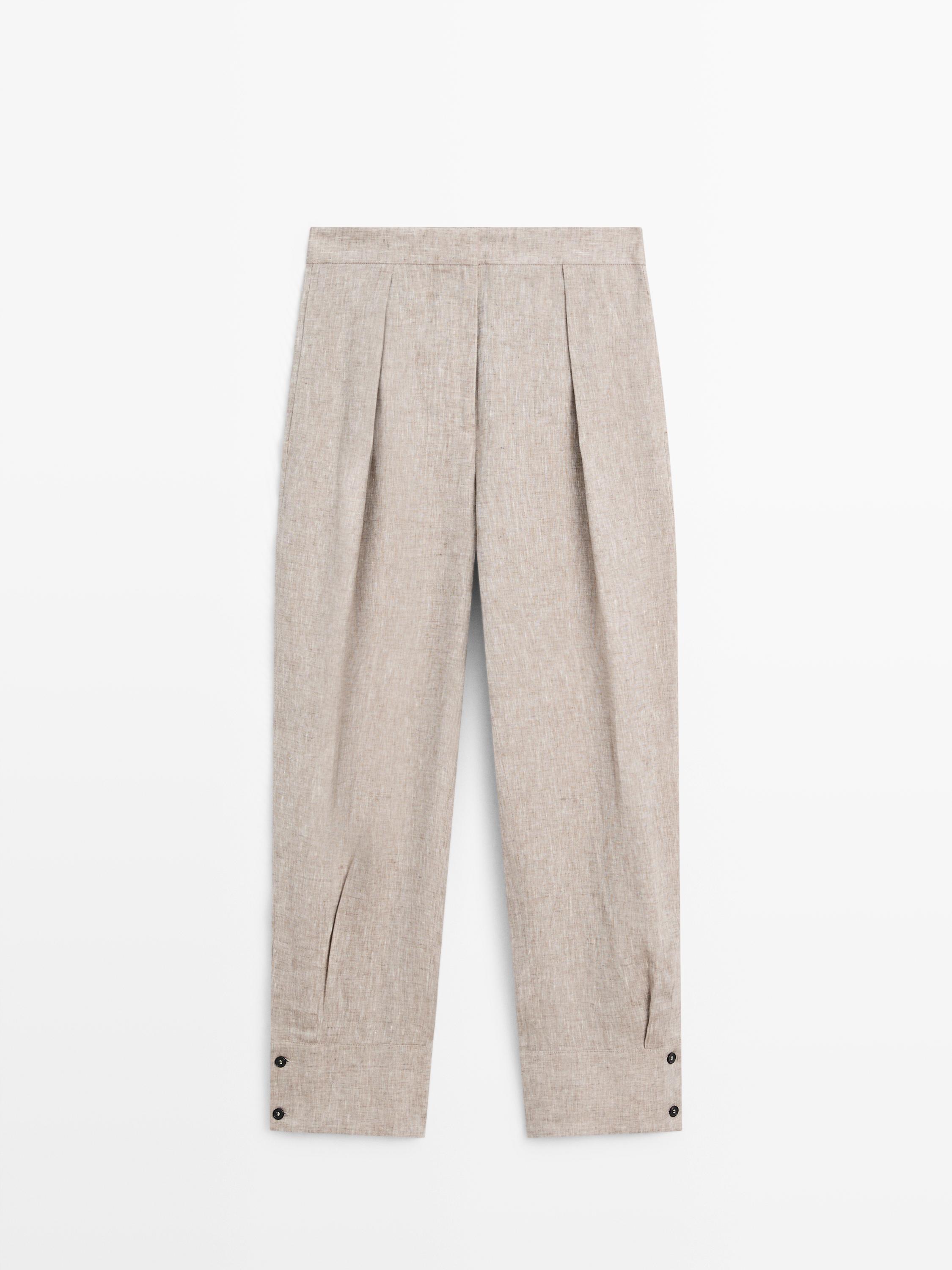 Вид спереди: бежевые брюки со складками, застежка на пуговицы по низу и деталь с пряжкой на талии.