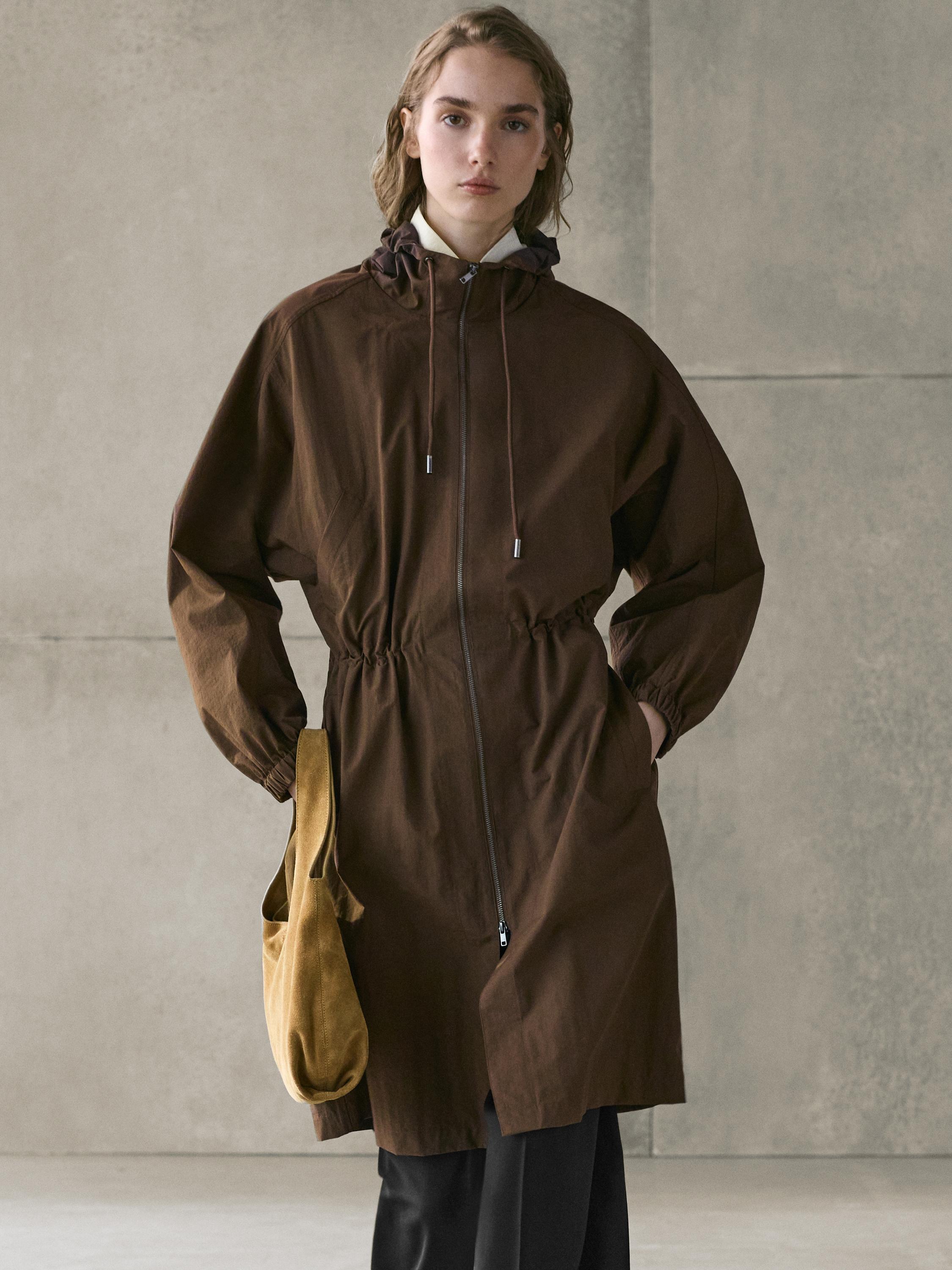Long brown parka set with hood and front zipper, black pants, and beige bag.