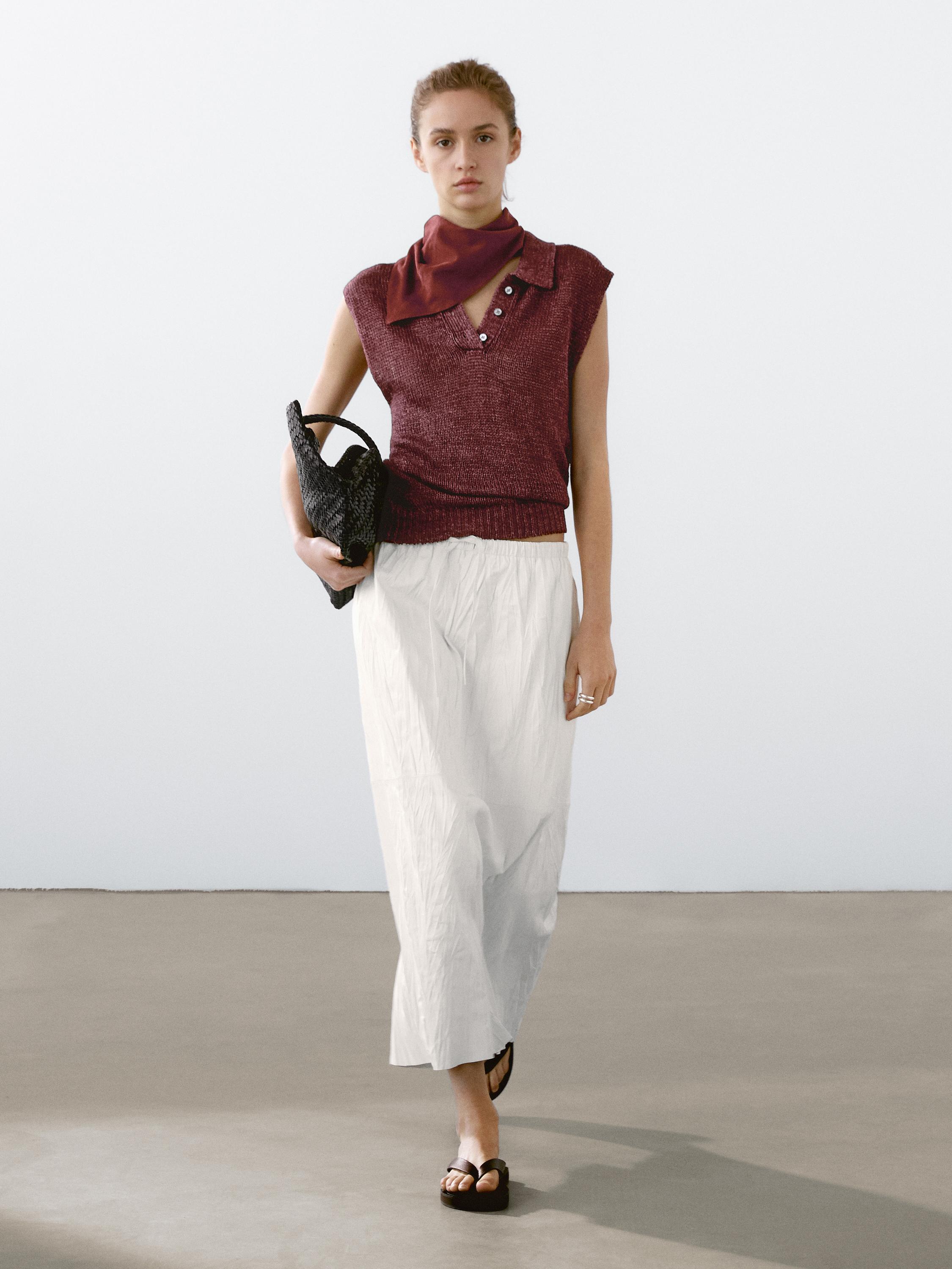 Ensemble composé d'un gilet bordeaux boutonné, d'une longue jupe blanche et d'une écharpe bordeaux, d'un sac noir en tissu et de sandales noires.