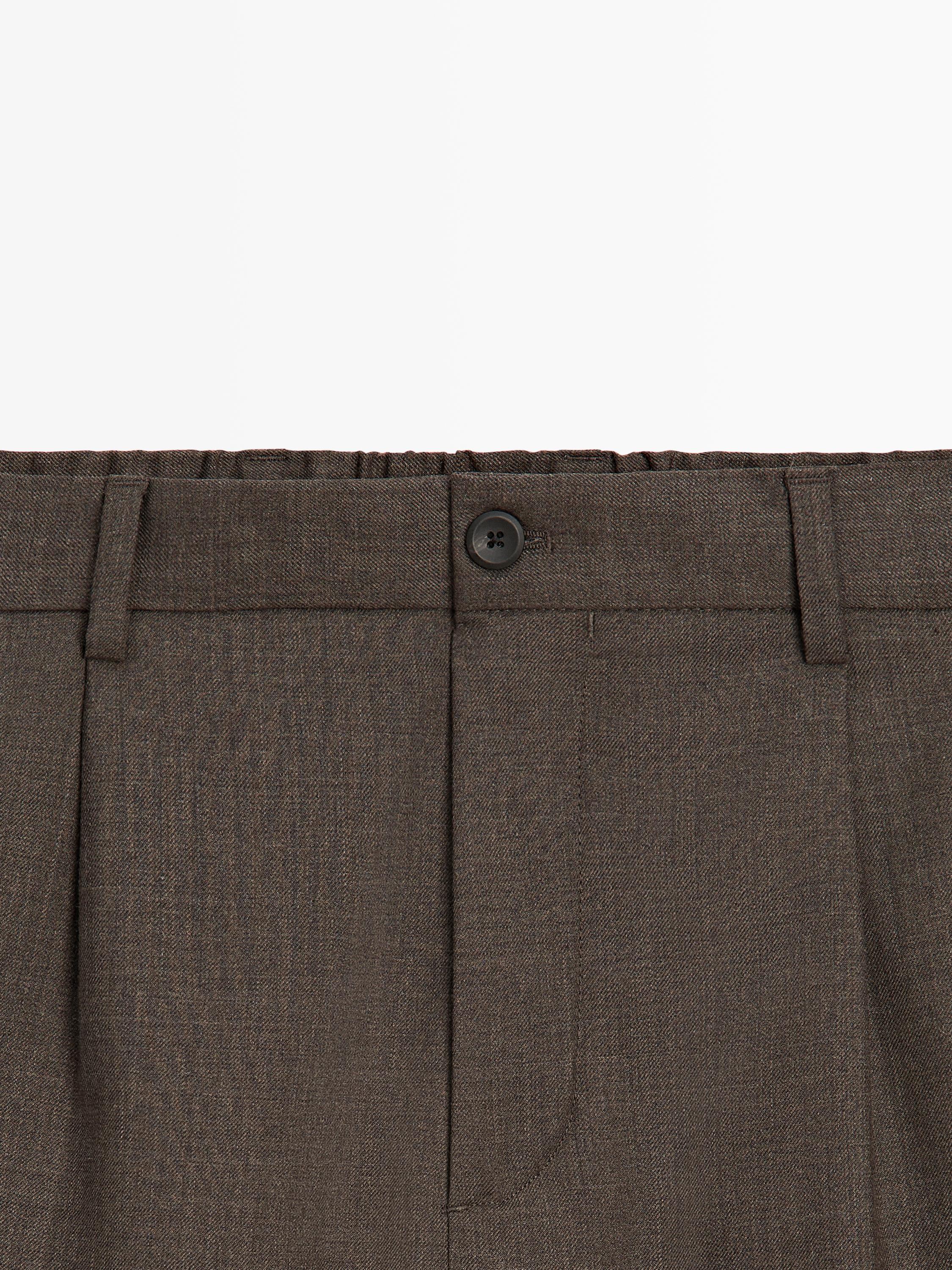 Plano médio da cintura de umas calças castanhas com pregas, botão preto e presilhas para cinto.