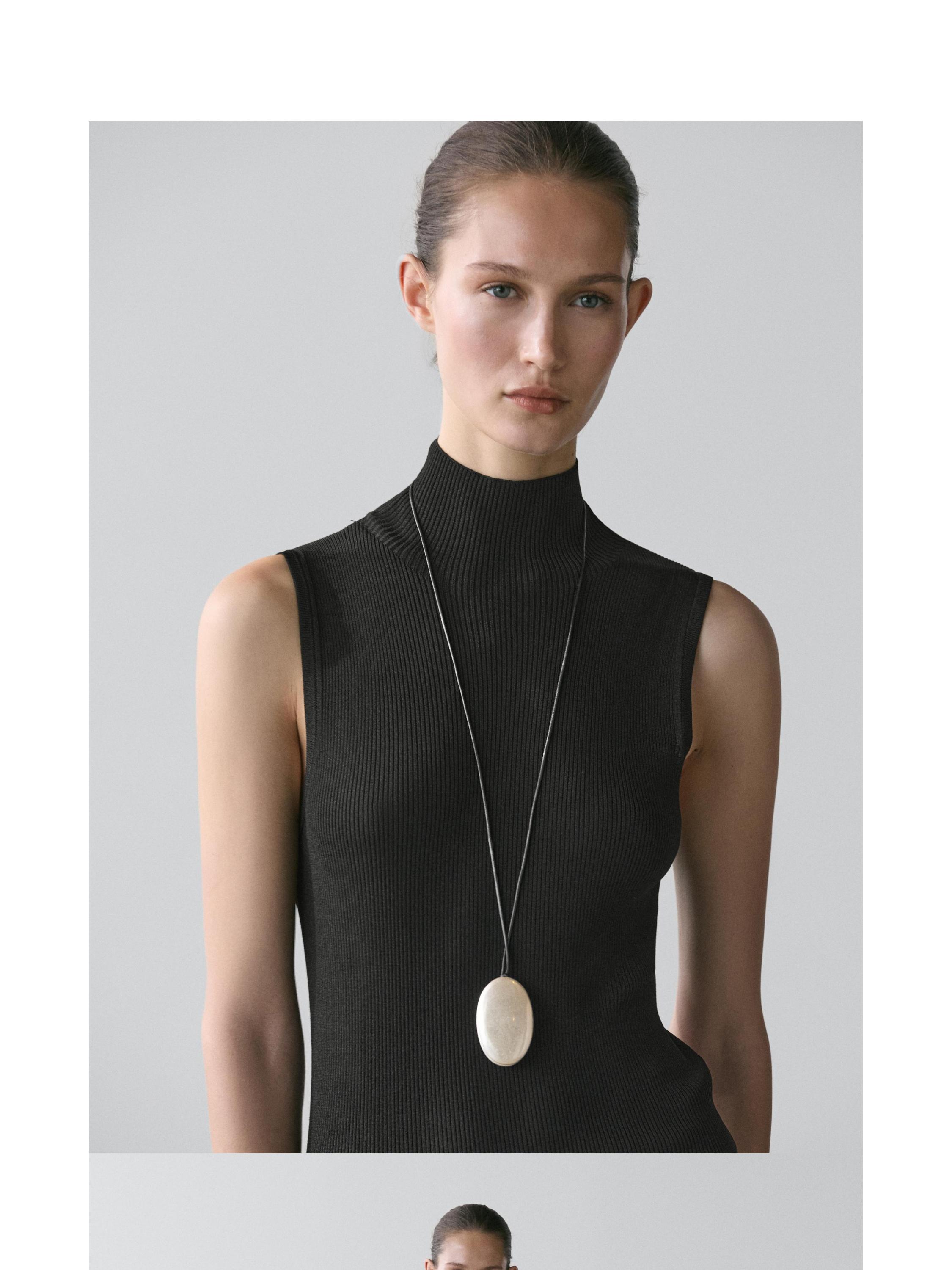 Plano medio de collar plateado con pieza ovalada sobre top negro sin mangas.