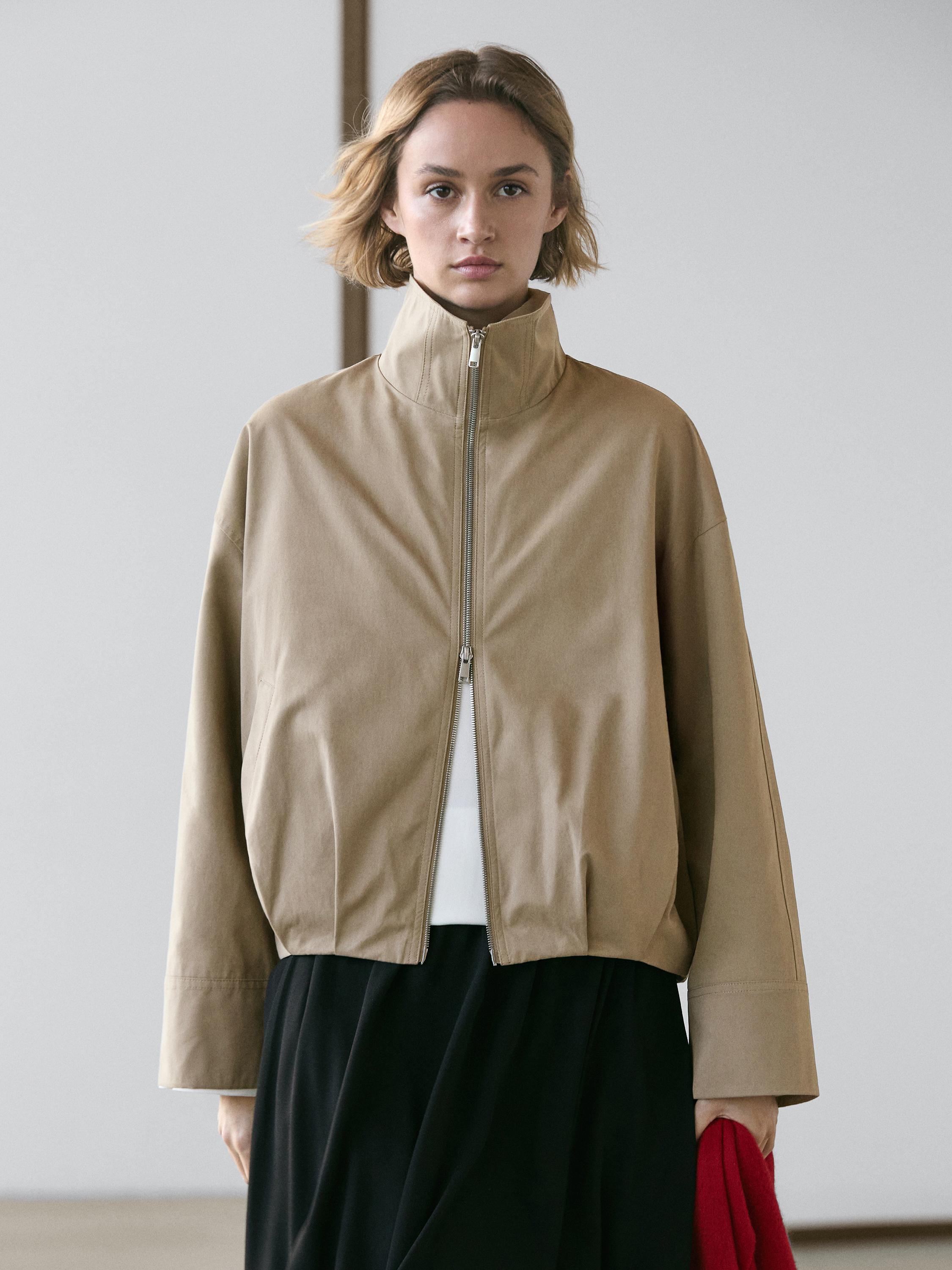 O'rta kadrda topo jigarrang bomber kurtka, old tarafida fermuar va baland yoqasi bilan.