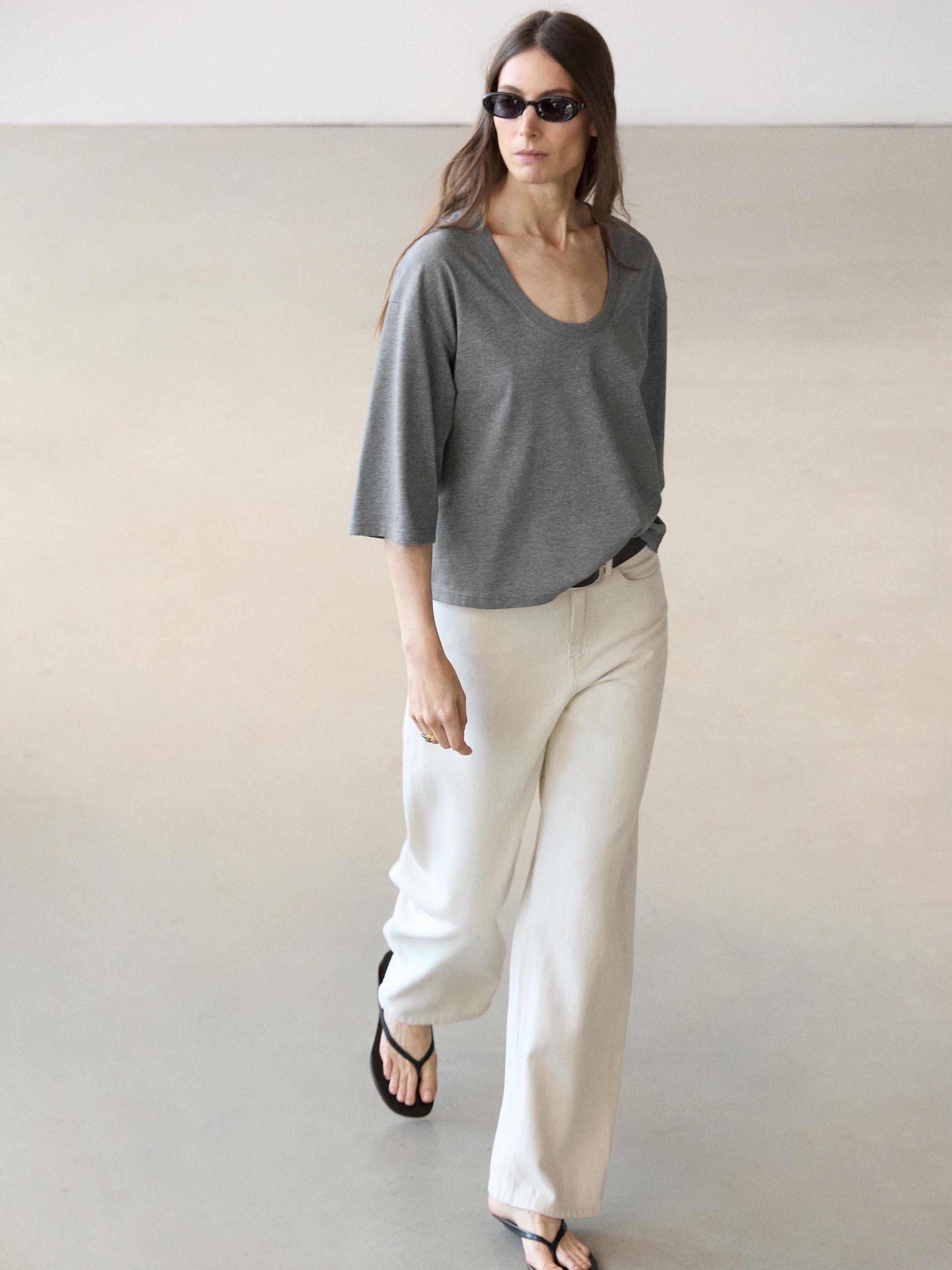 Graues Kurzarm-T-Shirt mit Rundhalsausschnitt und weiter weißer Hose sowie schwarzen Sandalen.