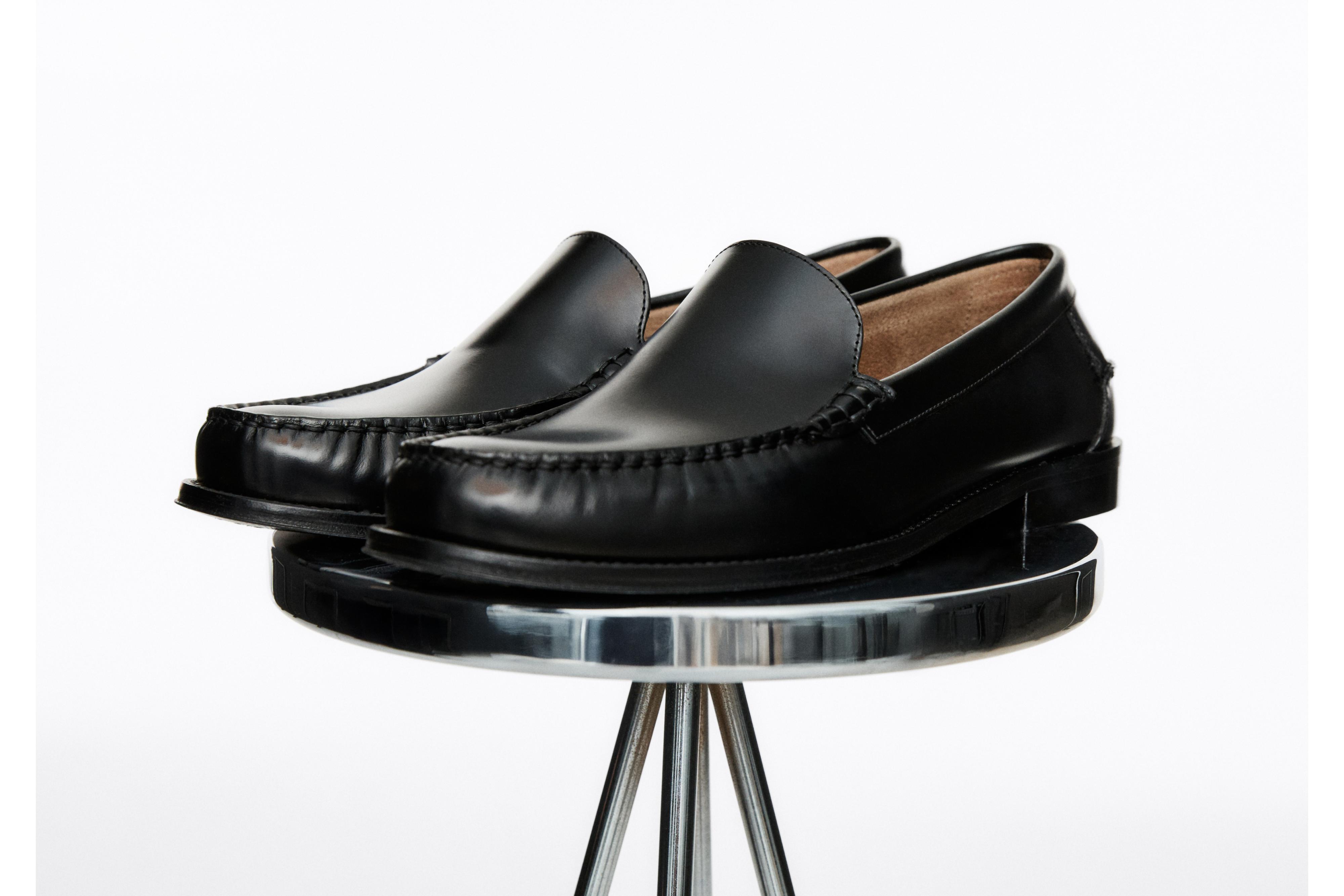 Këpucë të zeza me stil mokasine me qepje të dukshme dhe shollë të ulët, të paraqitura mbi një sipërfaqe metalike.
