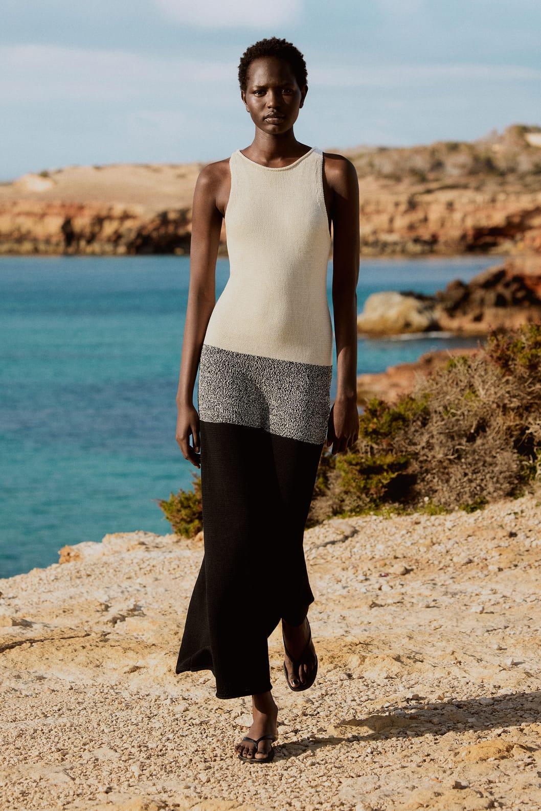 Modelo con vestido sin mangas beige y negro, caminando por la costa rocosa con el mar azul de fondo bajo un cielo despejado.