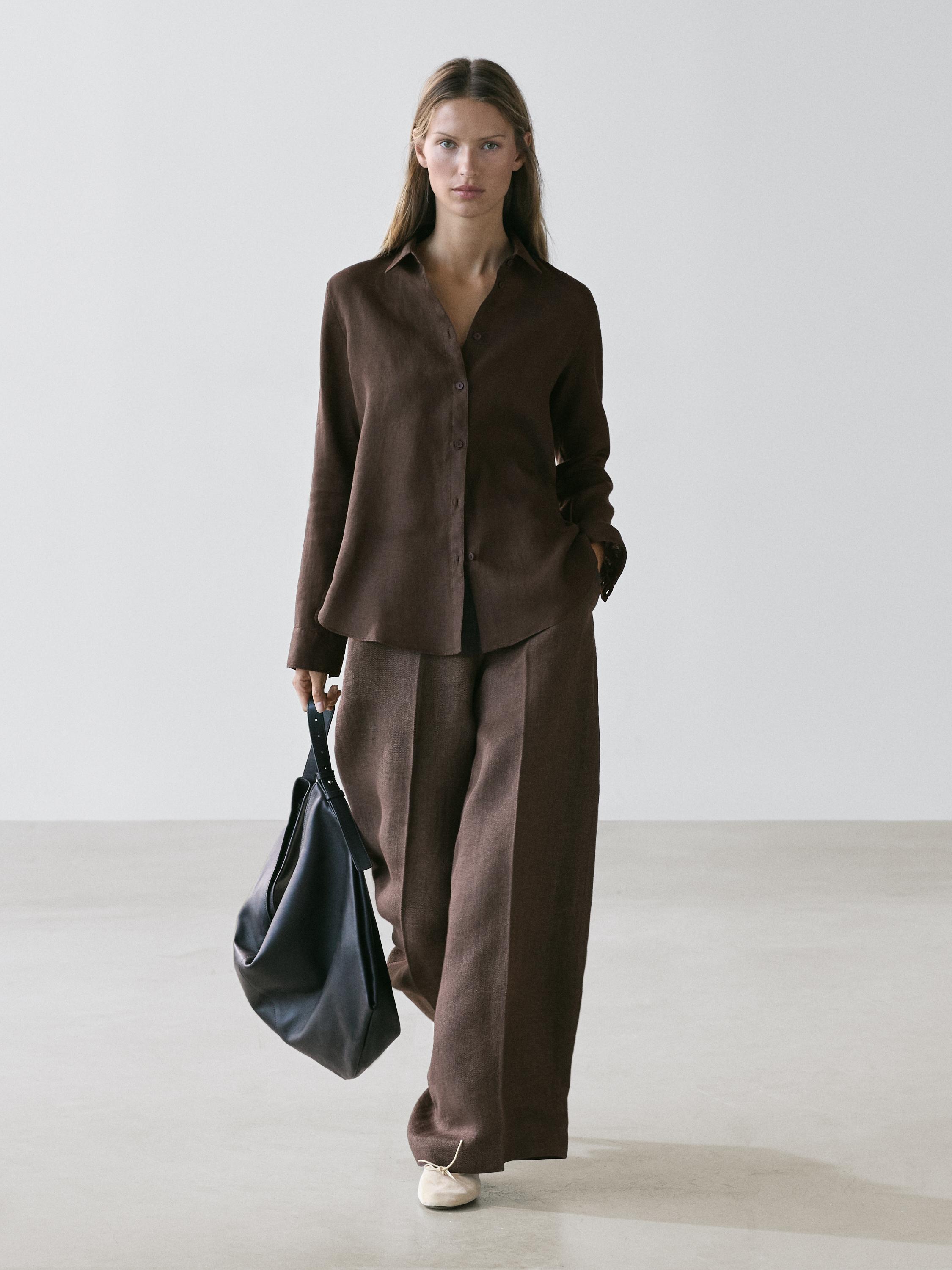 Комплект из коричневой рубашки на пуговицах и сочетающихся широких брюк. Носит черную сумку и светлую обувь.