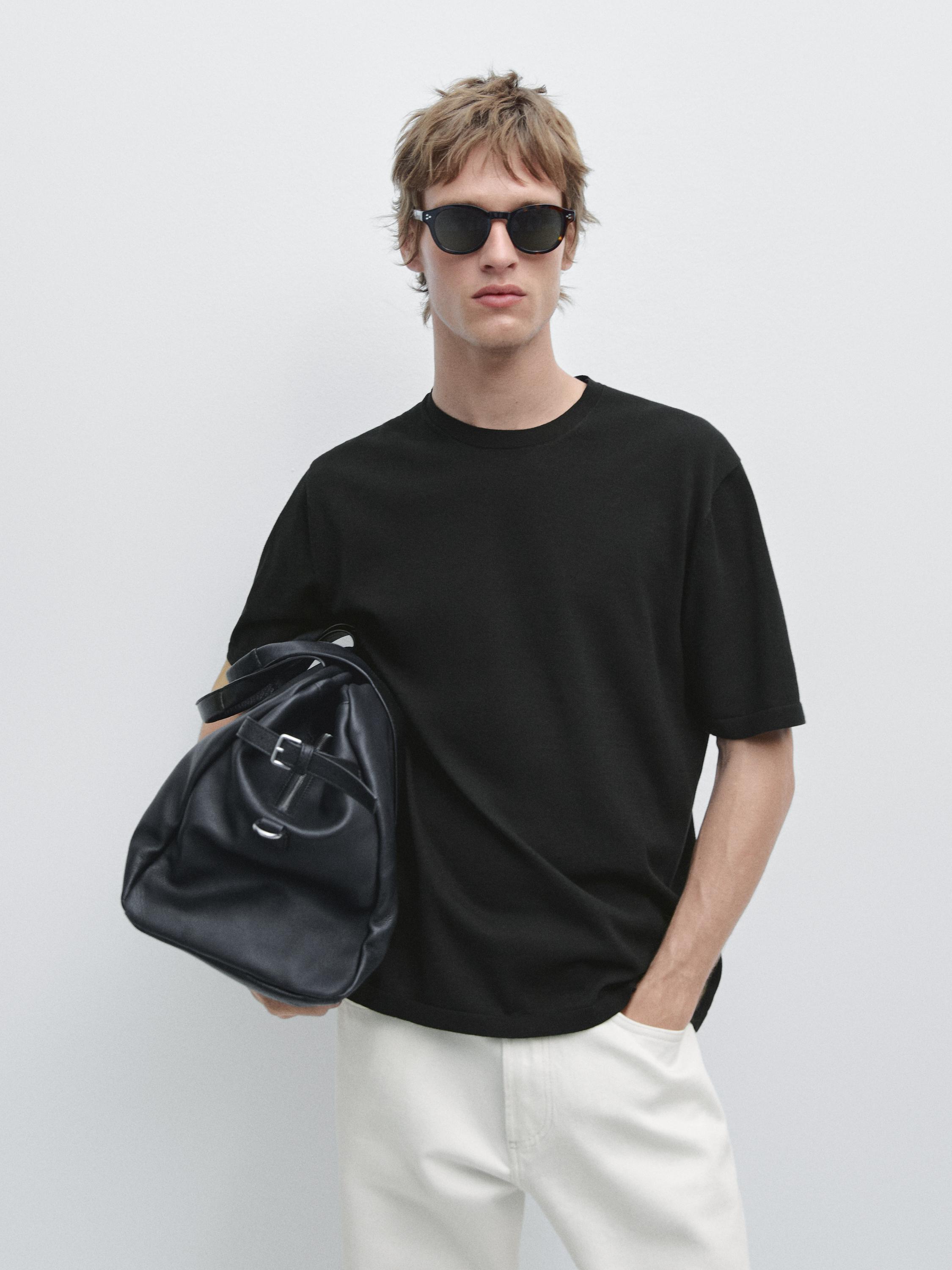 Plan moyen d'un T-shirt noir à manches courtes. Il porte un pantalon blanc et un sac noir au bras.