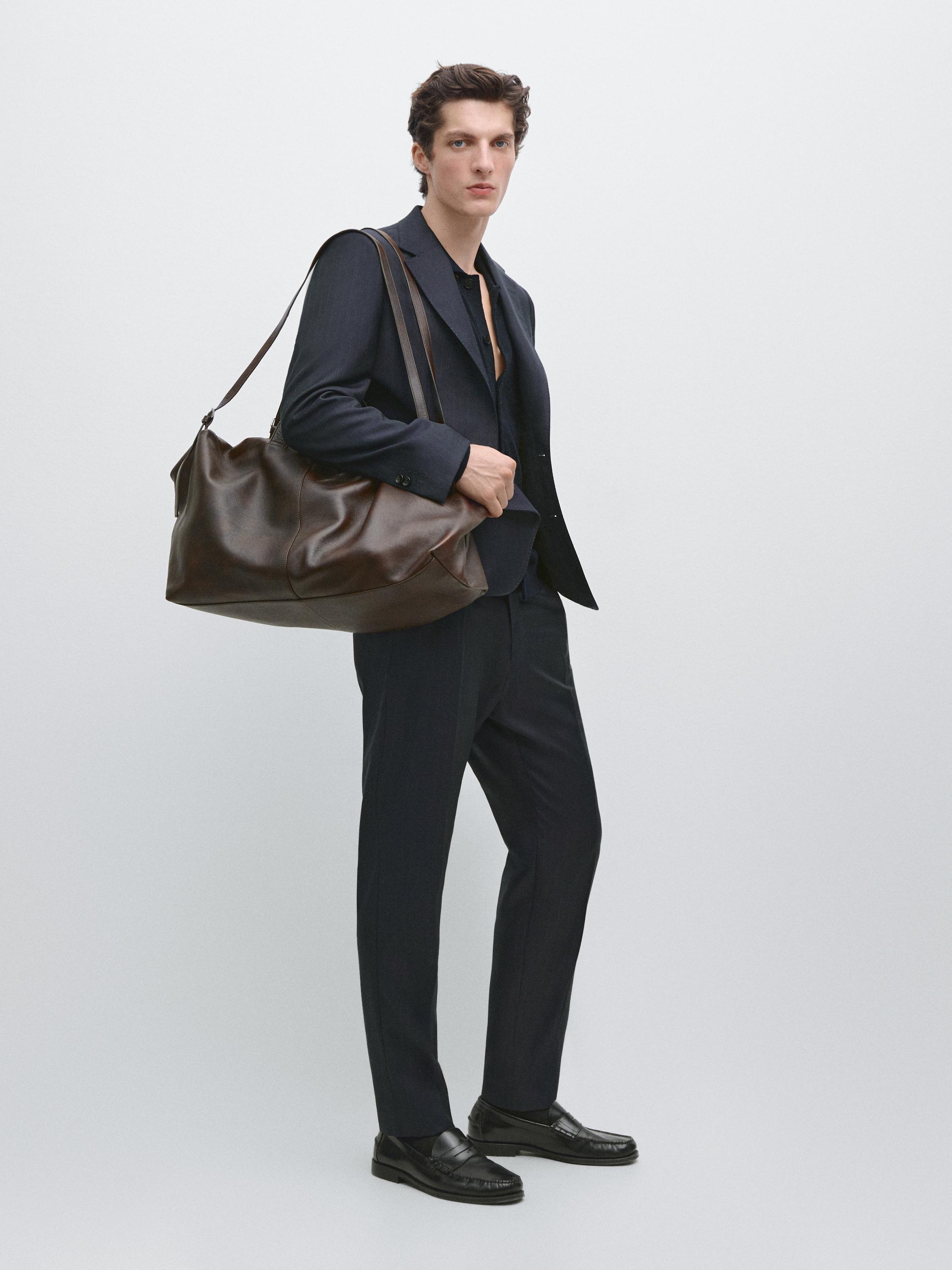 Conjunto de blazer azul-marinho com lapelas e calças a combinar, mala castanha grande e mocassins pretos.