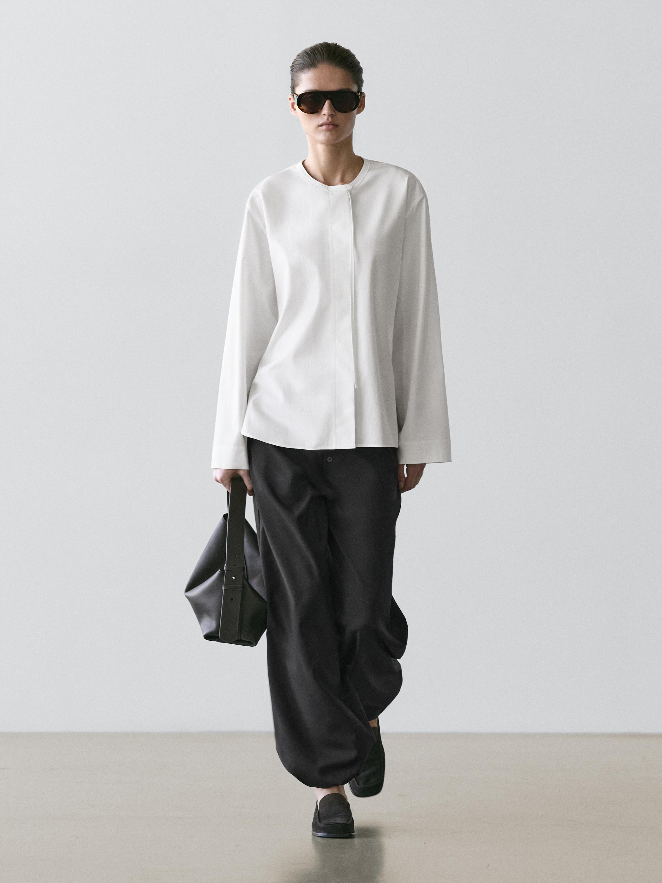 Ensemble composé d'une chemise blanche fluide à col rond et d'un pantalon noir ample. Il/Elle porte un sac noir et des mocassins.