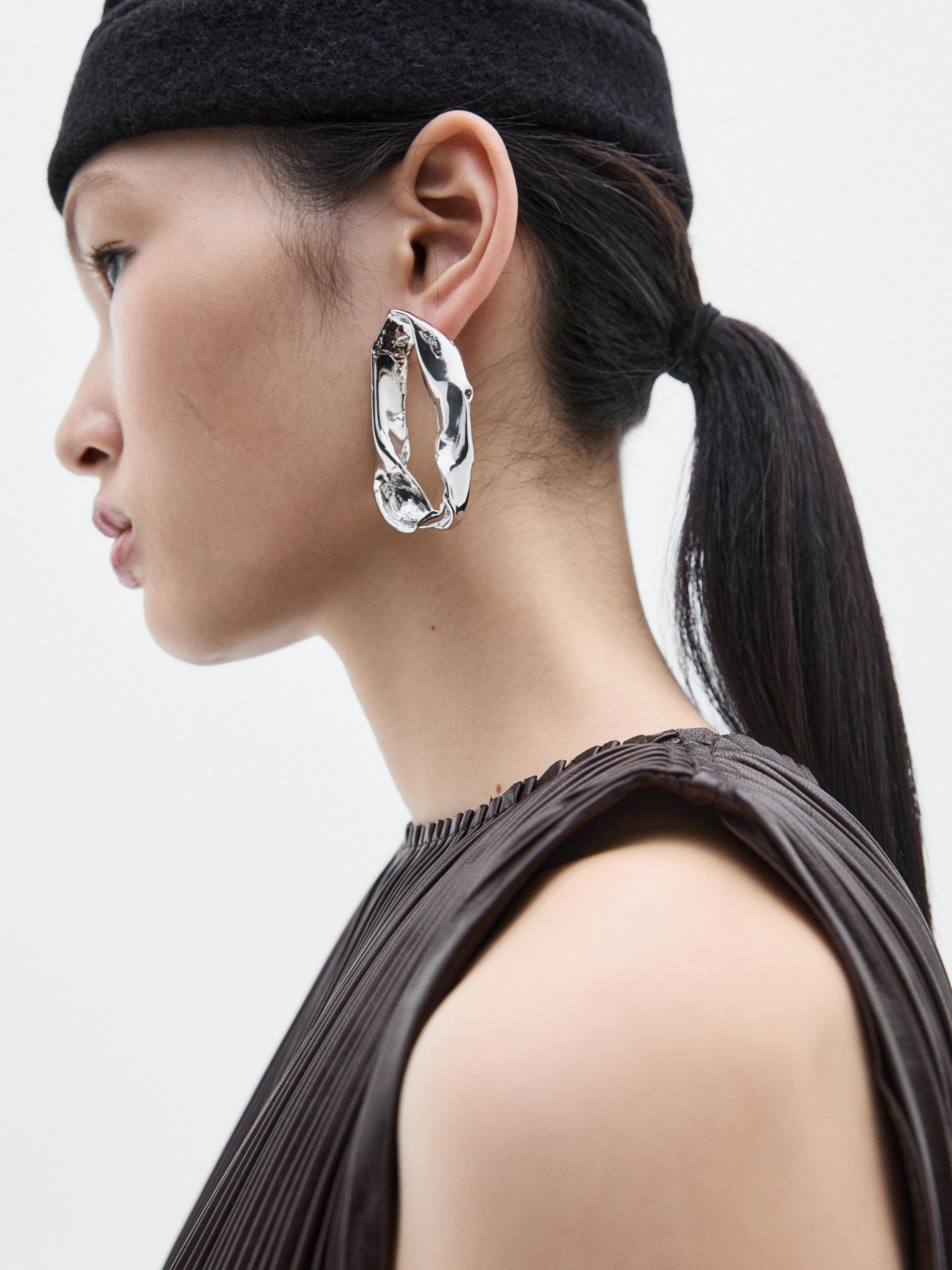 Primeiro plano de brinco prateado grande e blusa castanha plissada sem mangas.