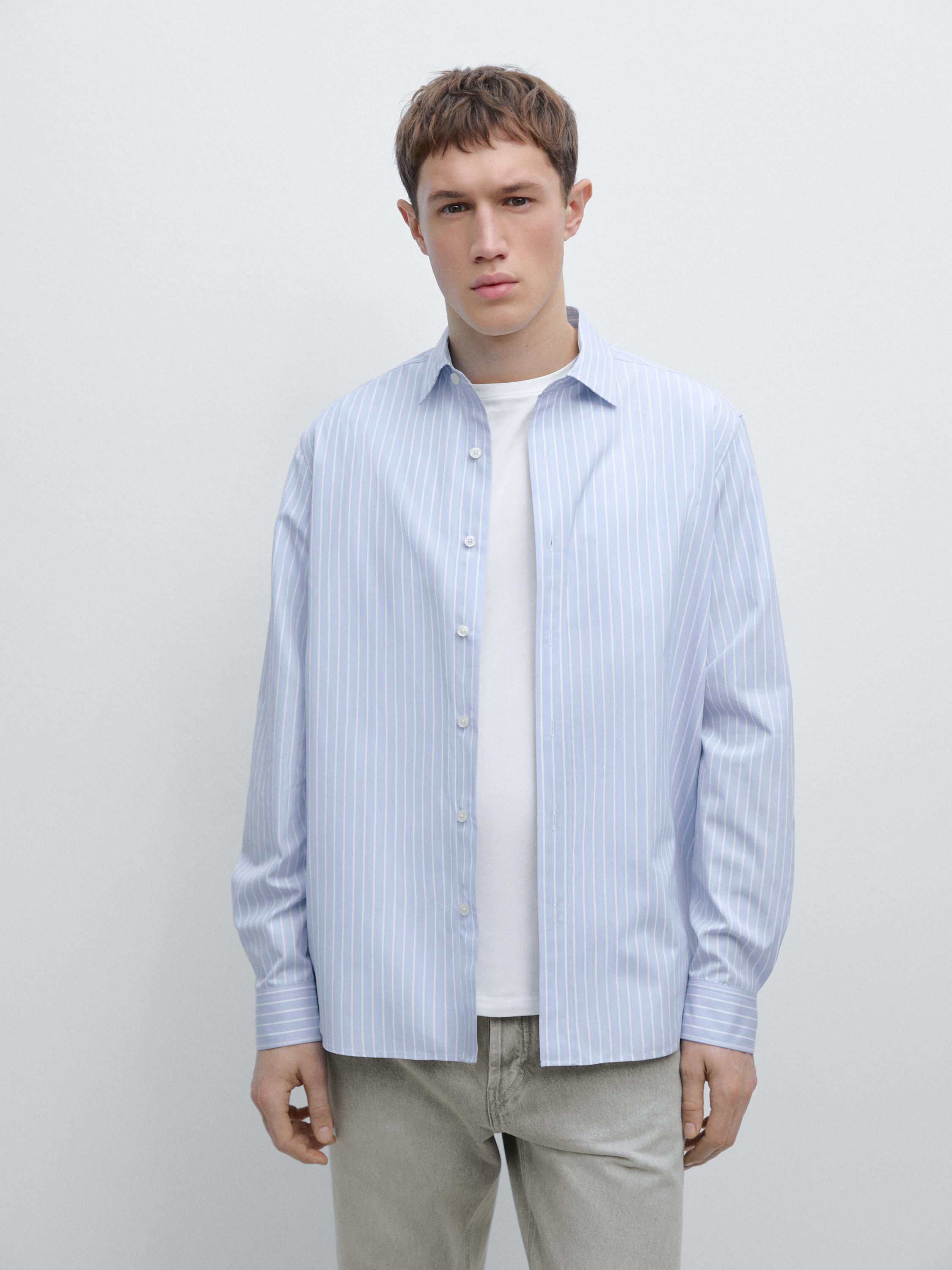 Zestaw składający się z błękitnej koszuli w pionowe pasy z długim rękawem, na białym podkoszulku, ze spodniami w kolorze szarym.