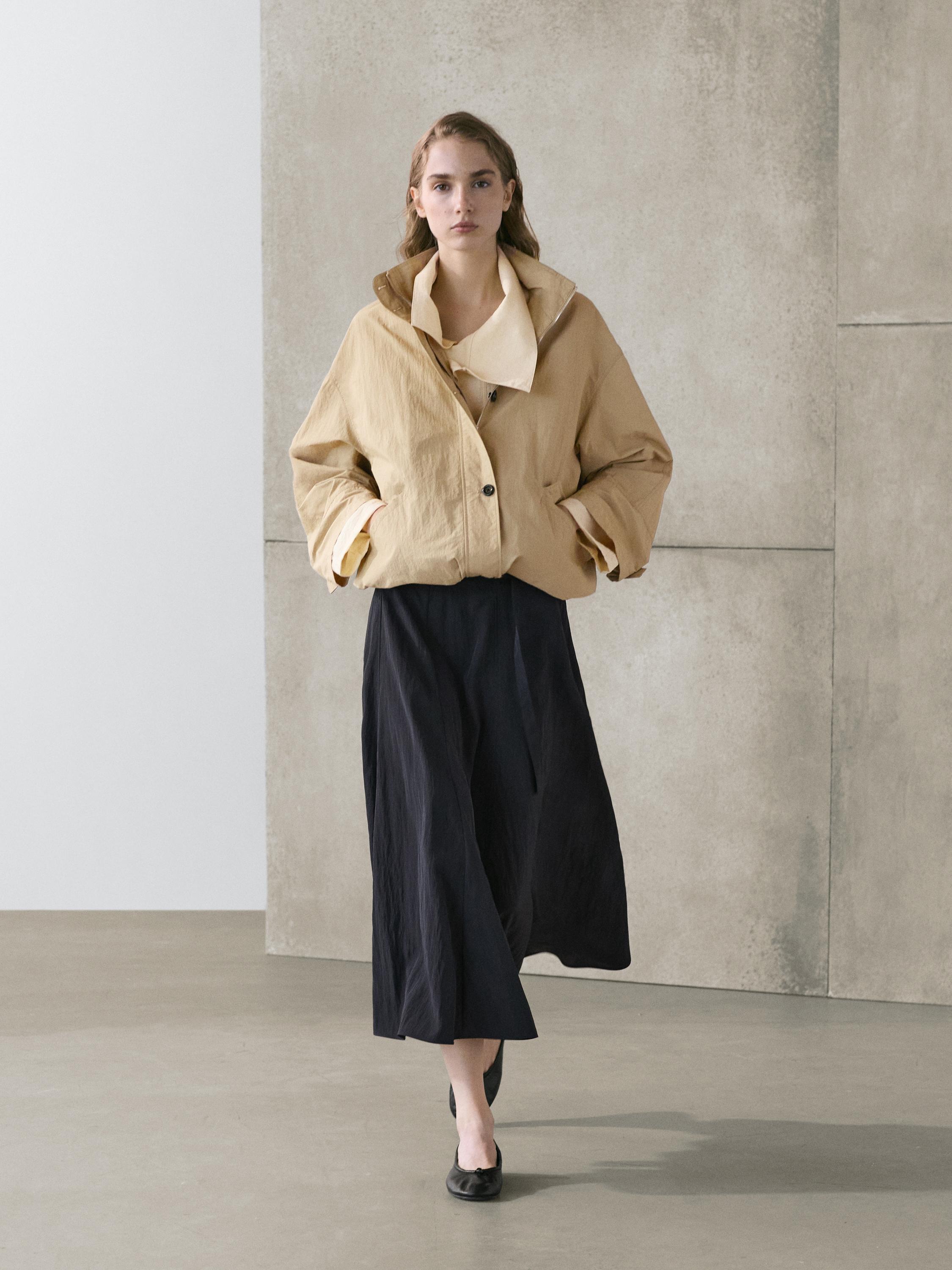 Zestaw składający się z długiej spódnicy w kolorze szaro-niebieskim z paskiem i beżowej marynarki z bocznymi kieszeniami i guzikami z przodu.