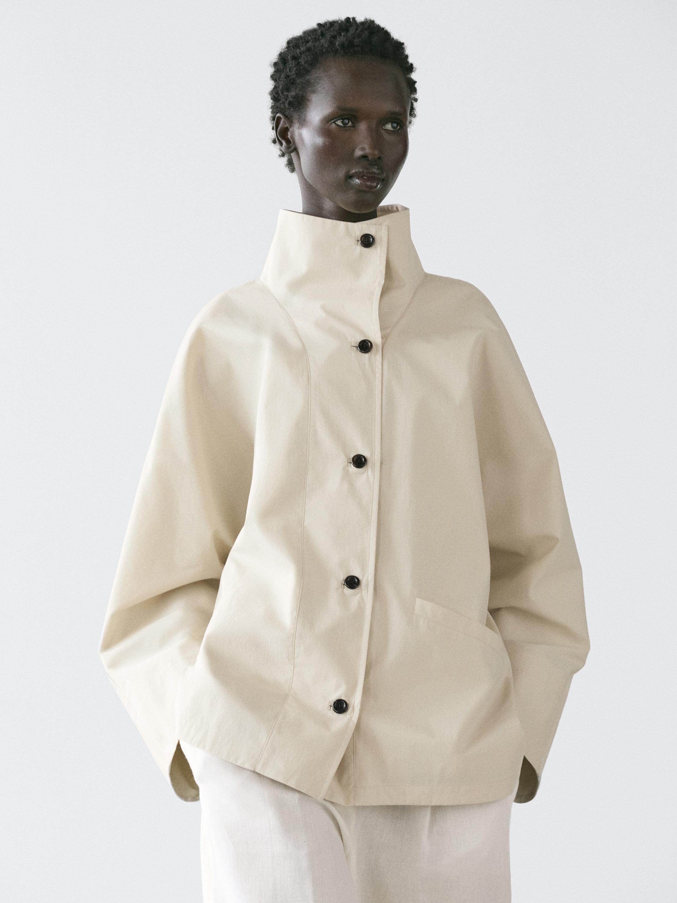Комплект из необработанной куртки с воротником-стойкой и черными пуговицами, боковыми карманами, поверх светлых брюк.