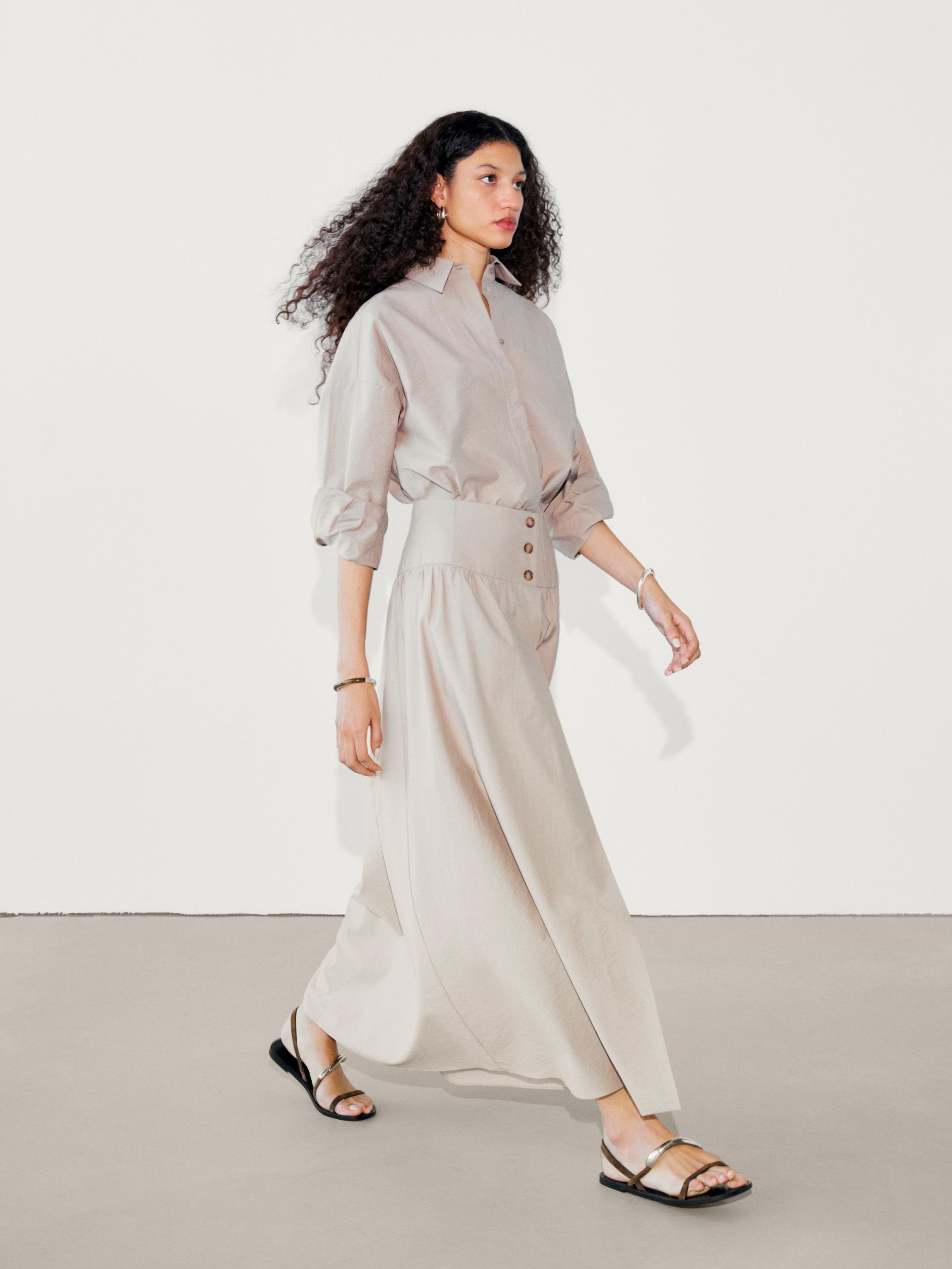 Ensemble composé d'un chemisier beige à manches longues et d'une jupe longue beige boutonnée sur le devant. Sandales noires visibles.