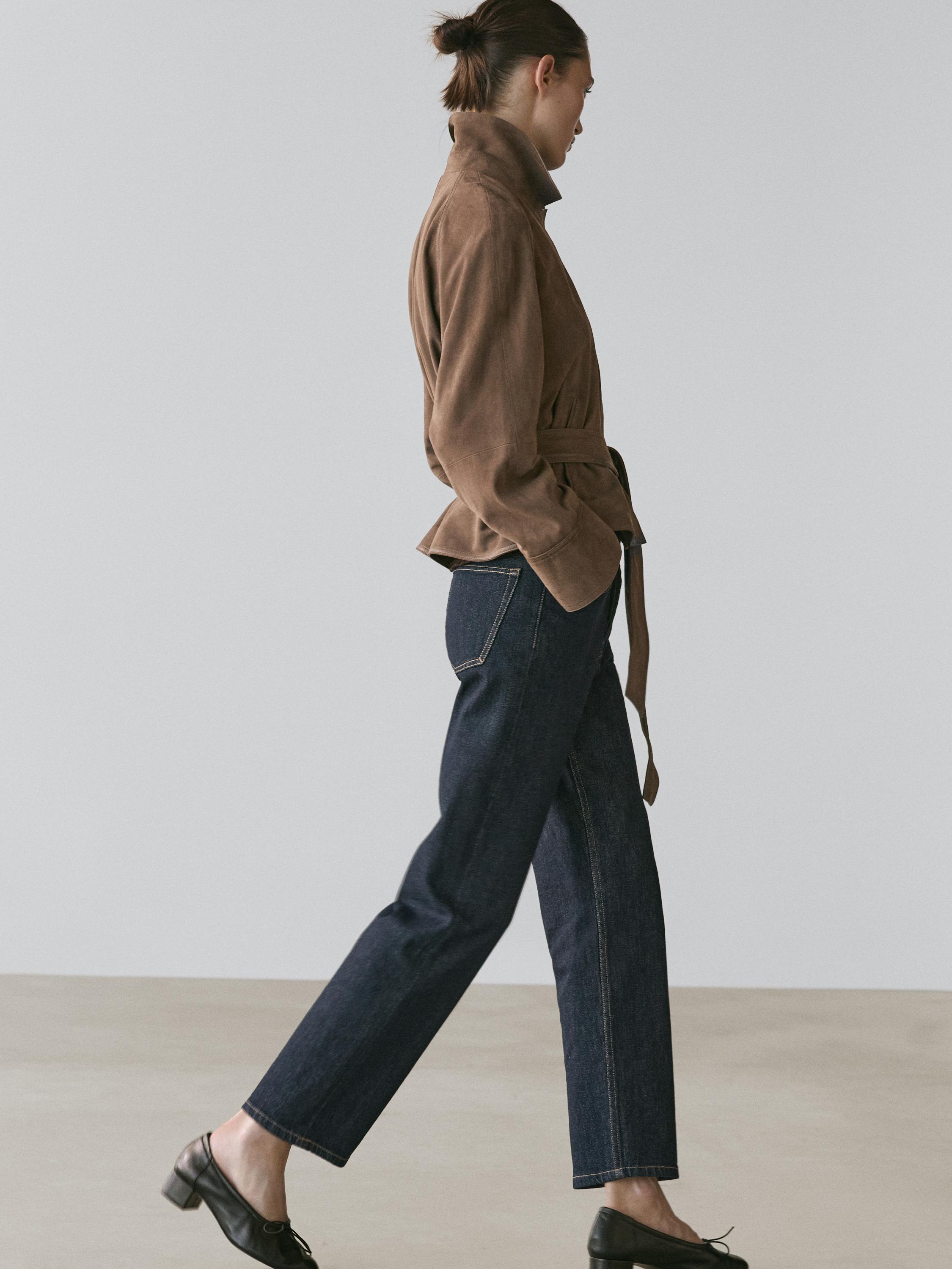 Conjunto de chaqueta bomber marrón topo con cinturón y jeans azul oscuro con bolsillos traseros visibles.