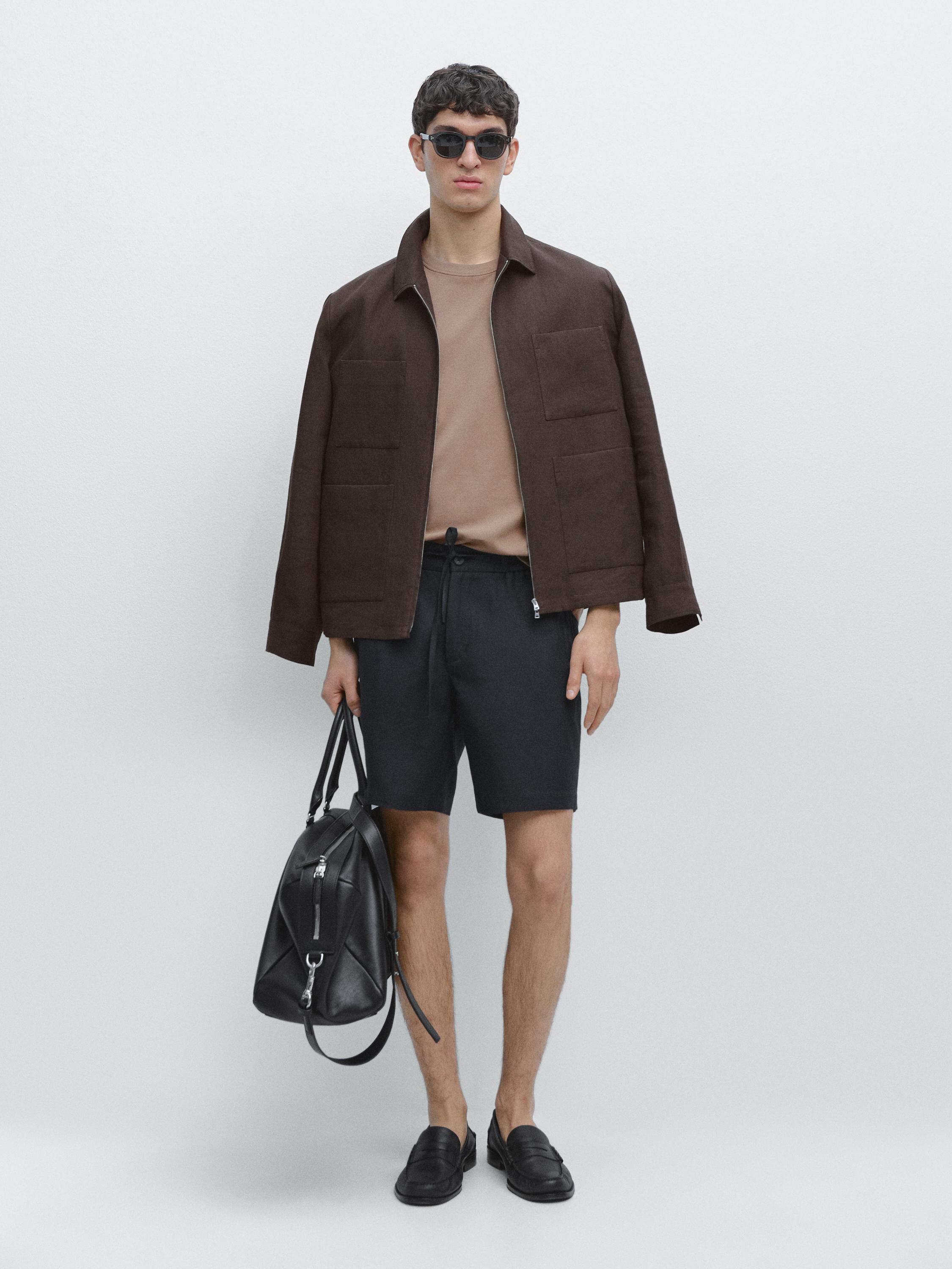 Conjunto de chamarra café con bolsillos, playera beige, bermudas azul marino y mocasines negros. Lleva bolsa negra.