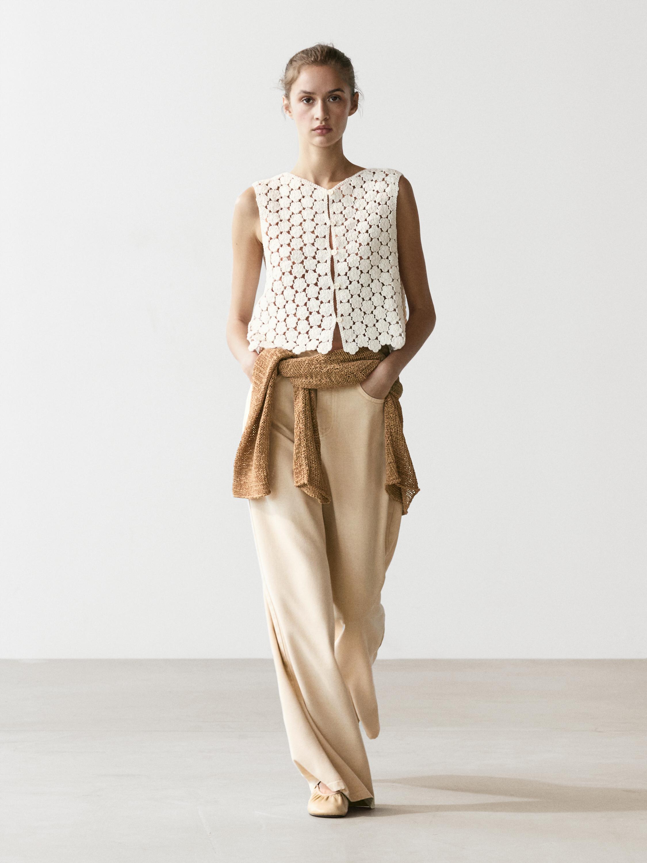 Conjunto de chaleco crochet crudo con detalle de flores, pantalones beige anchos y suéter marrón atado a la cintura.