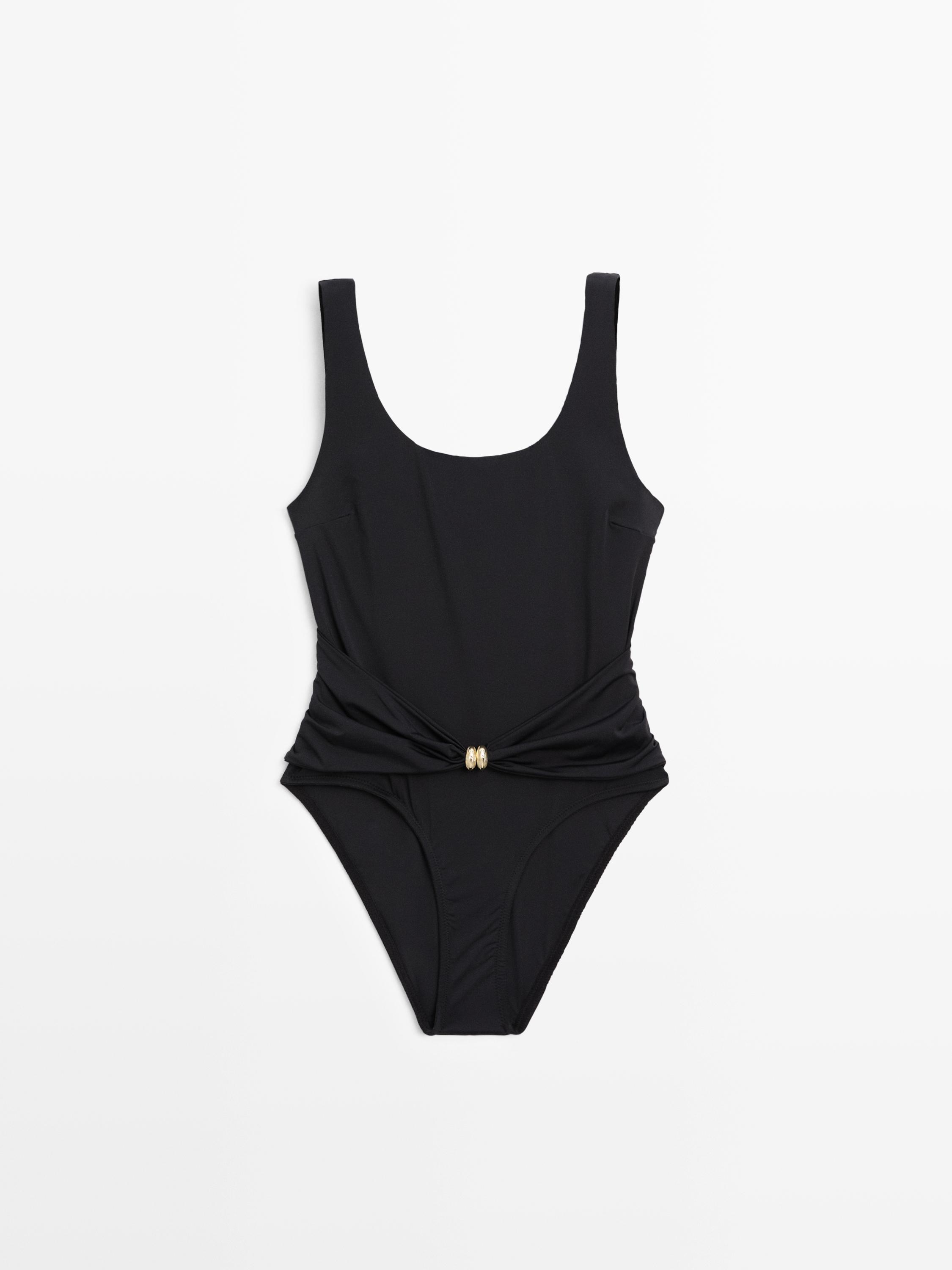 Vista frontal de traje de baño negro con detalle de joya dorada en la cintura y tirantes anchos.