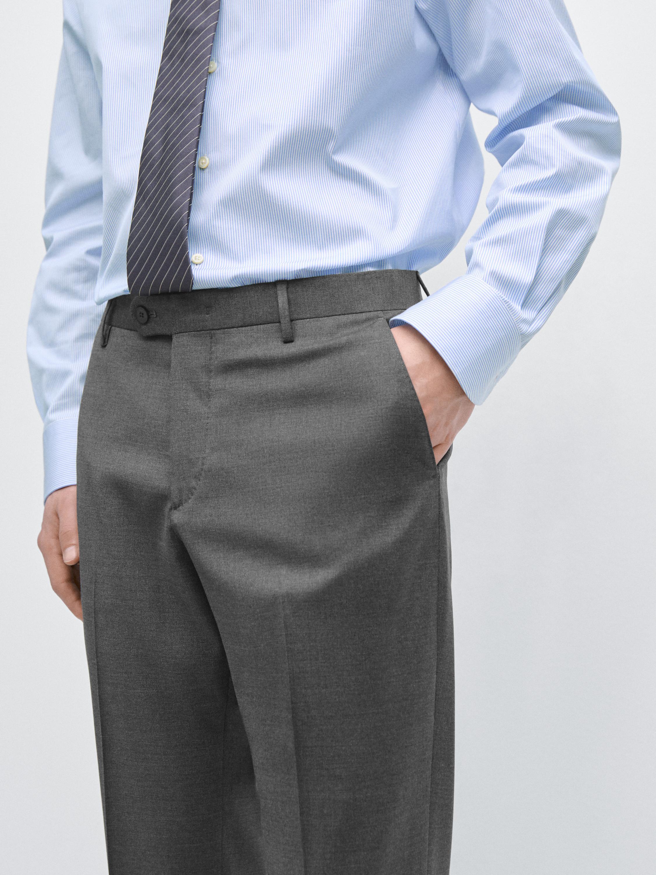 Medium shot van een grijze kostuumbroek met steekzakken en een tailleband met riemlussen. Een blauw overhemd en een stropdas zijn zichtbaar.
