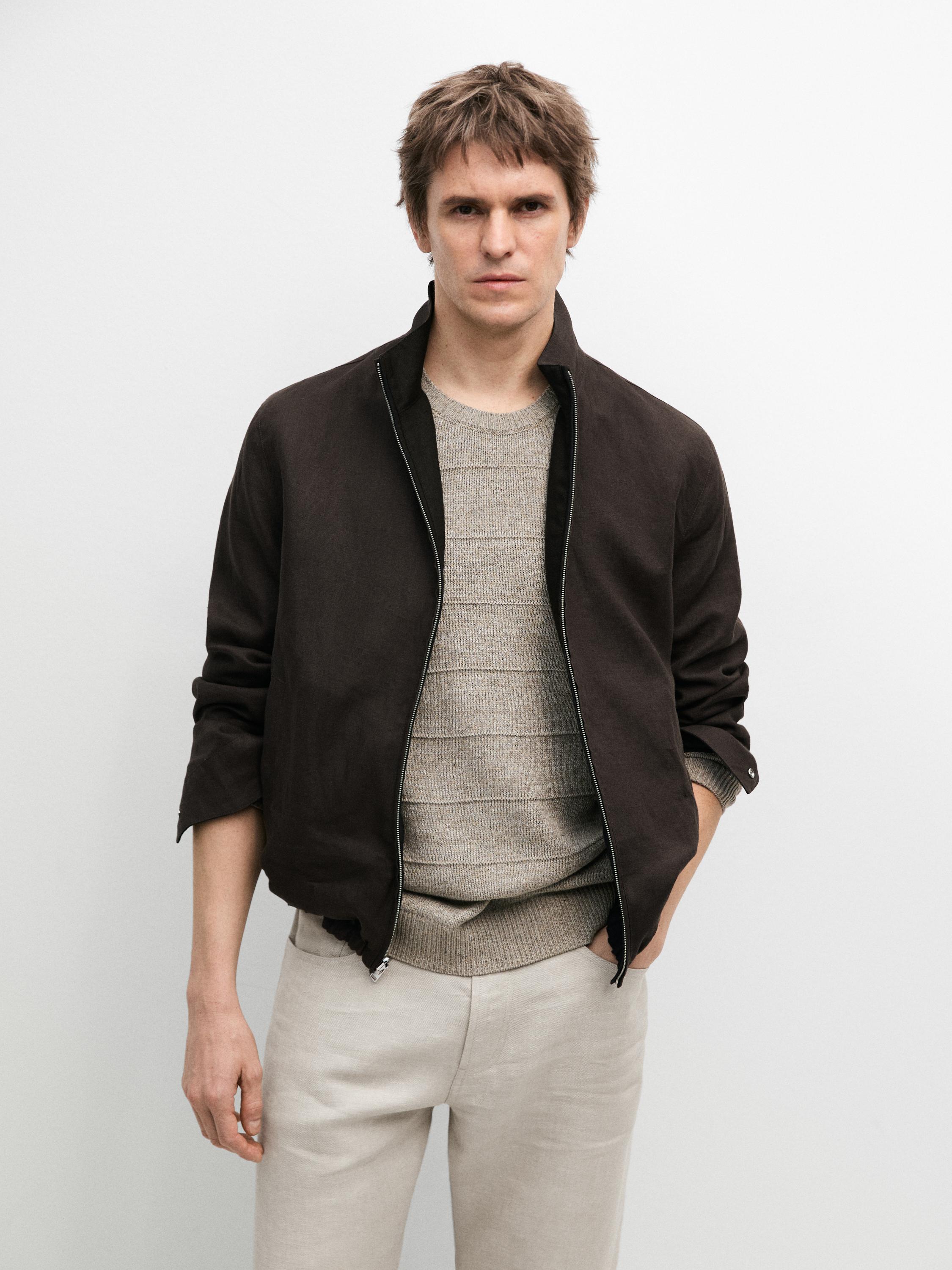 Schwarze Jacke mit Reißverschluss und gestreifter beigefarbener Pullover, helle Hose.