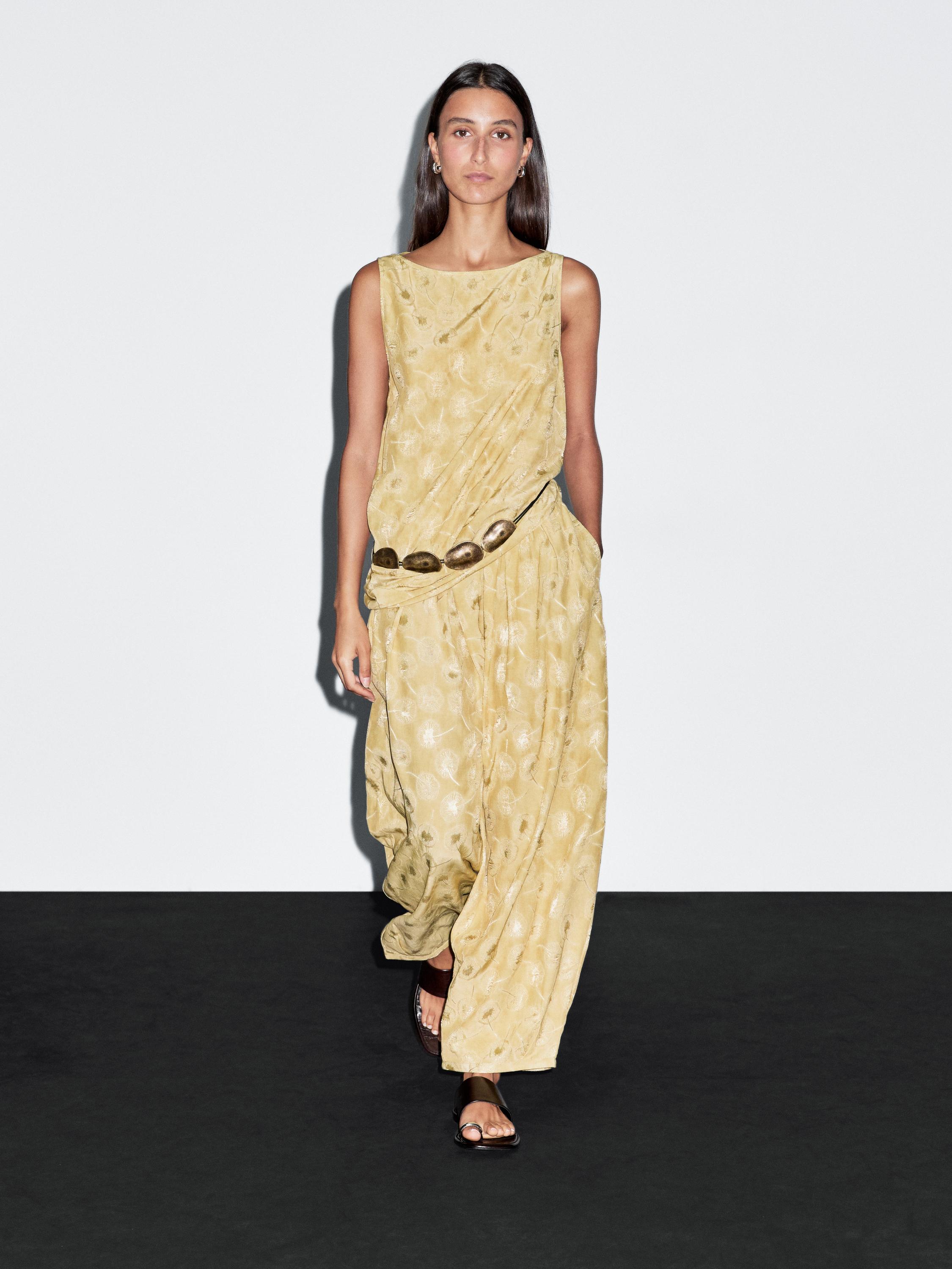 Mustard yellow maxi dress set with abstract print, decorative belt with metallic details, and flat sandals.