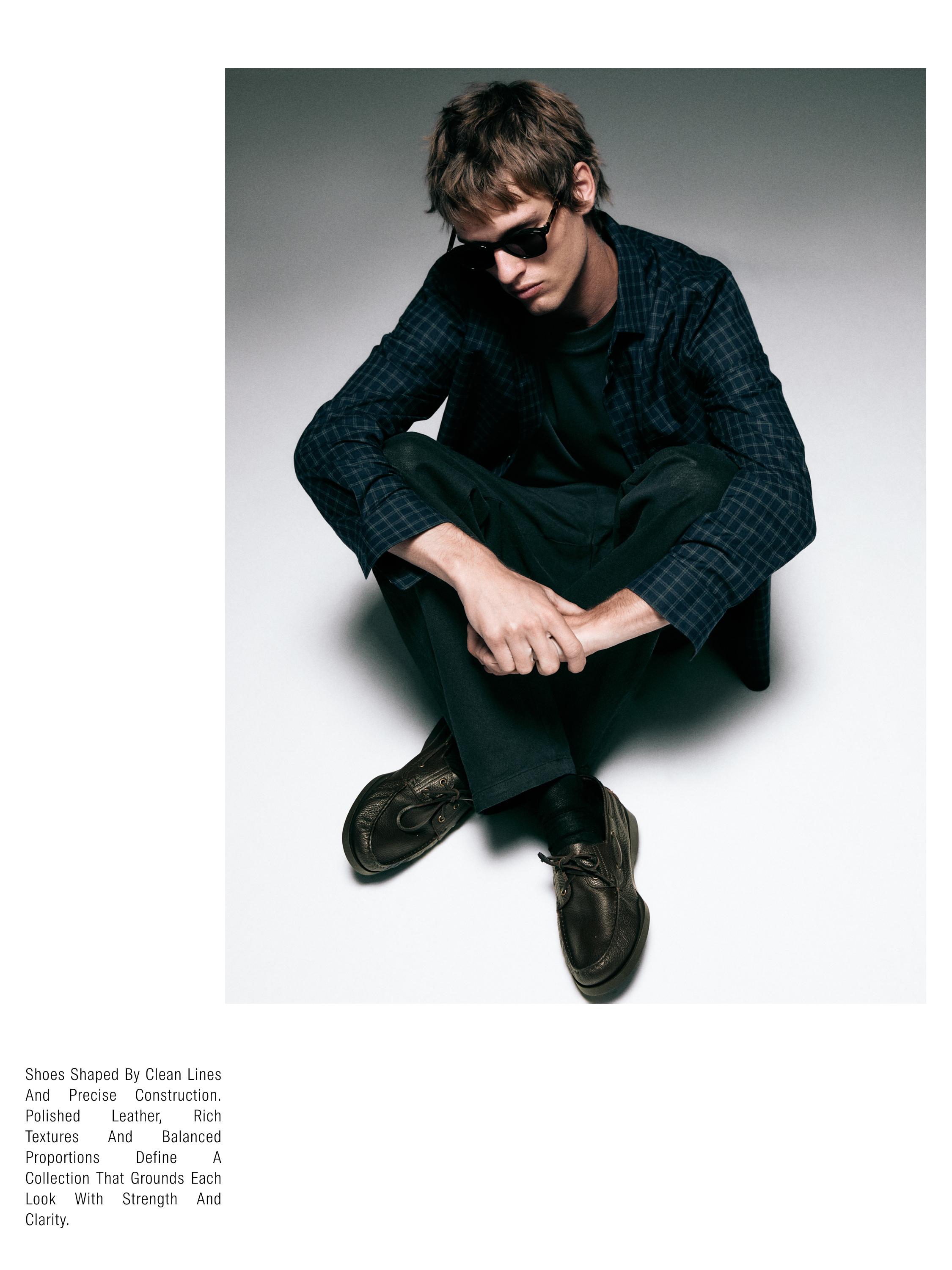 Komplet me këmishë me kuadrate blu të errët, bluzë të zezë dhe pantallona të errëta. Këpucë kafe të dukshme.