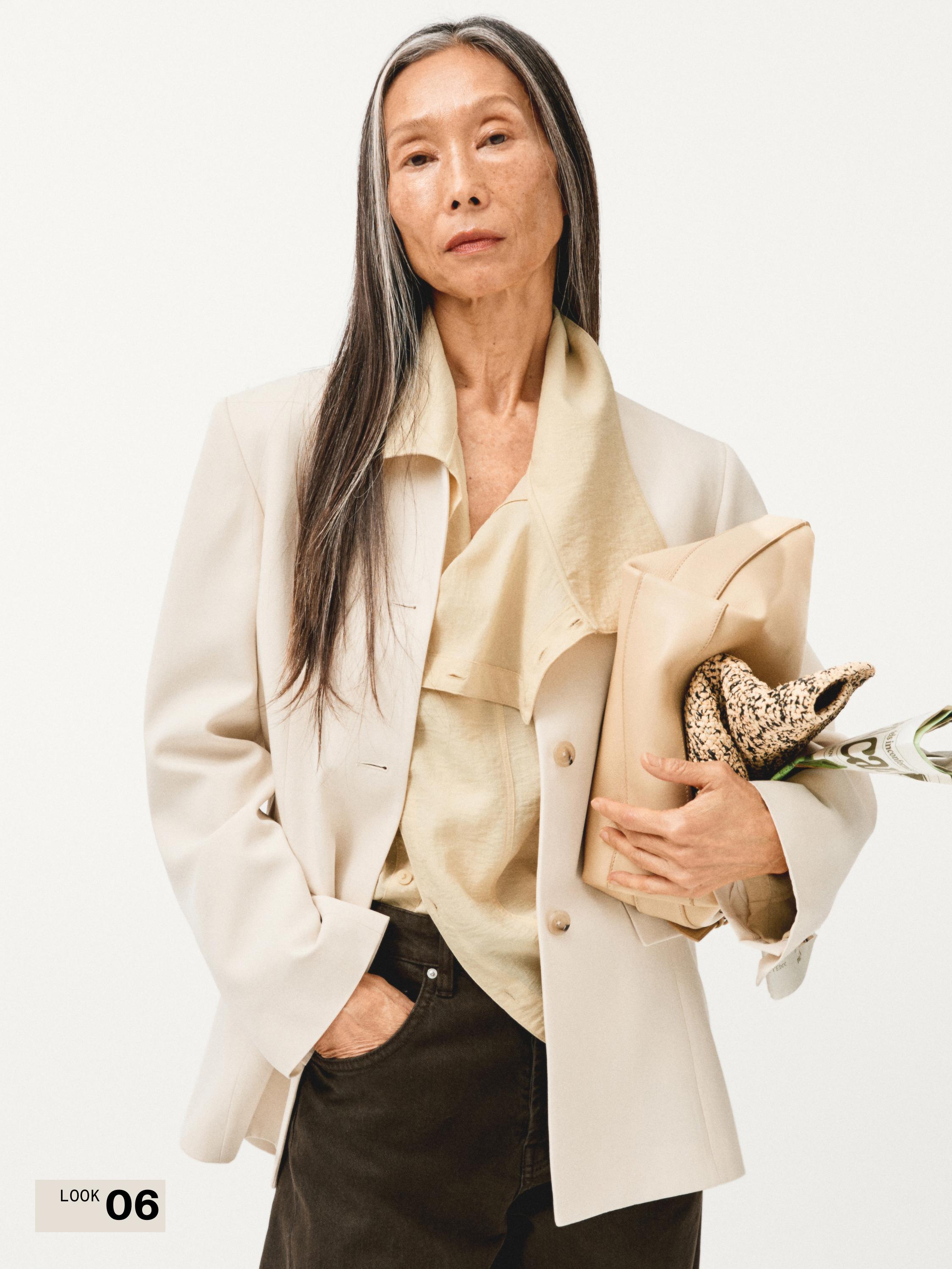 طقم بليزر بيج، بلوزة كريمية بياقة واسعة وبنطال داكن. أزرار ظاهرة واليد في الجيب.