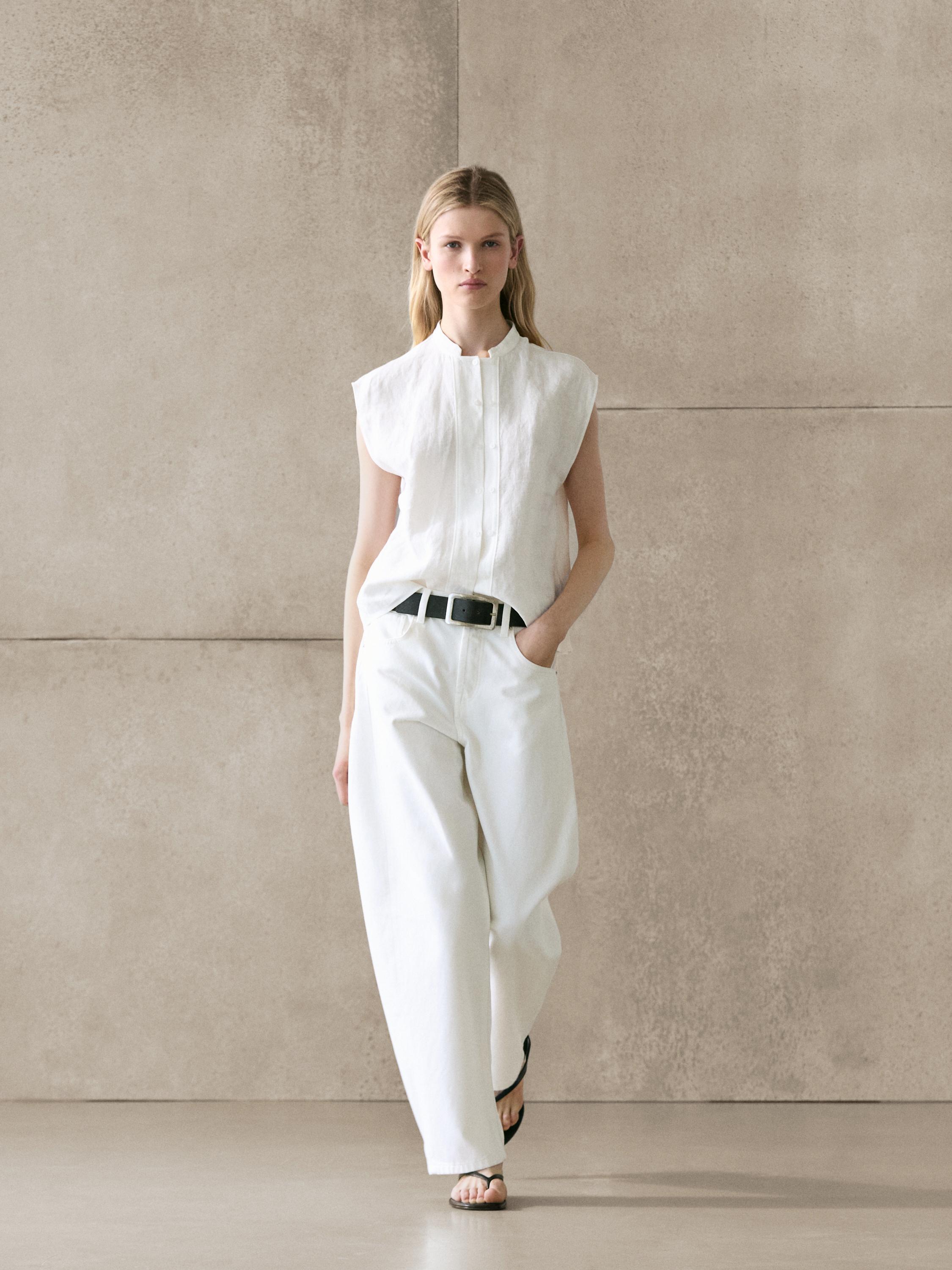 Conjunto de camisa blanca sin mangas con botones y pantalones anchos blancos con cinturón negro.