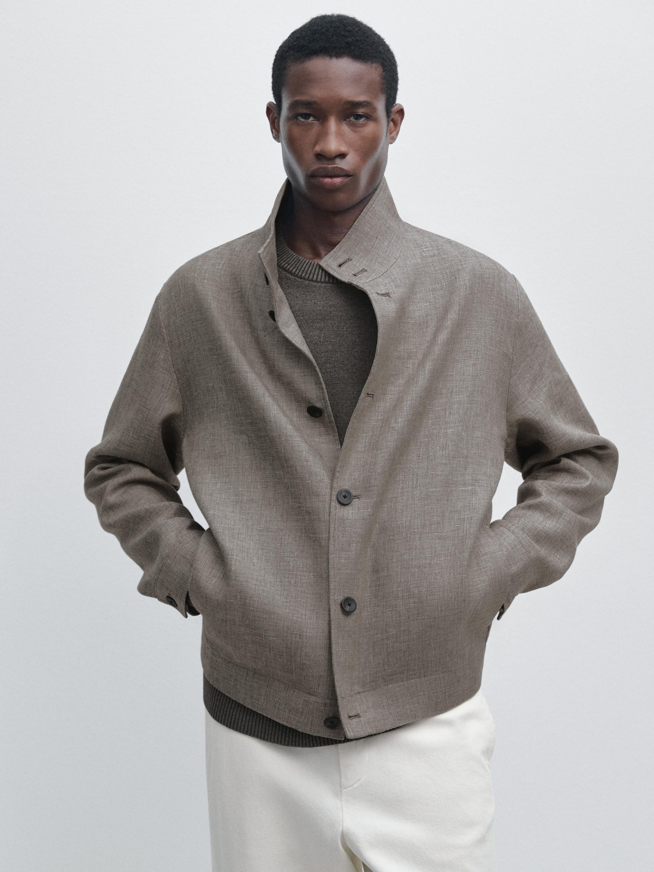 Комплект: бежева сорочка-куртка з темними ґудзиками та бічними кишенями, поверх коричневого светра та світлих штанів.