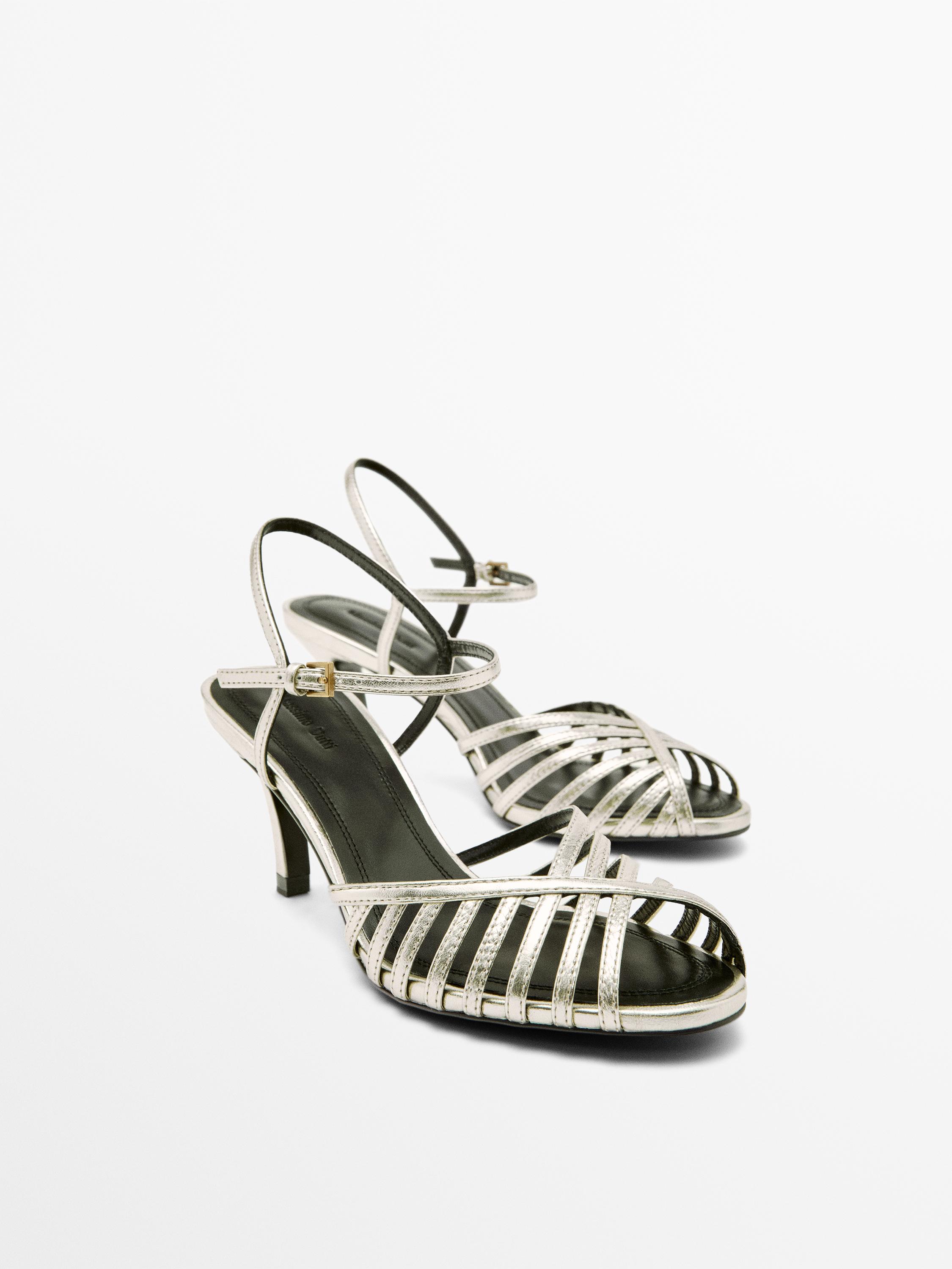 صندل بكعب عالٍ بلون البلاتين مع أشرطة رفيعة متقاطعة وإبزيم إغلاق عند الكاحل.