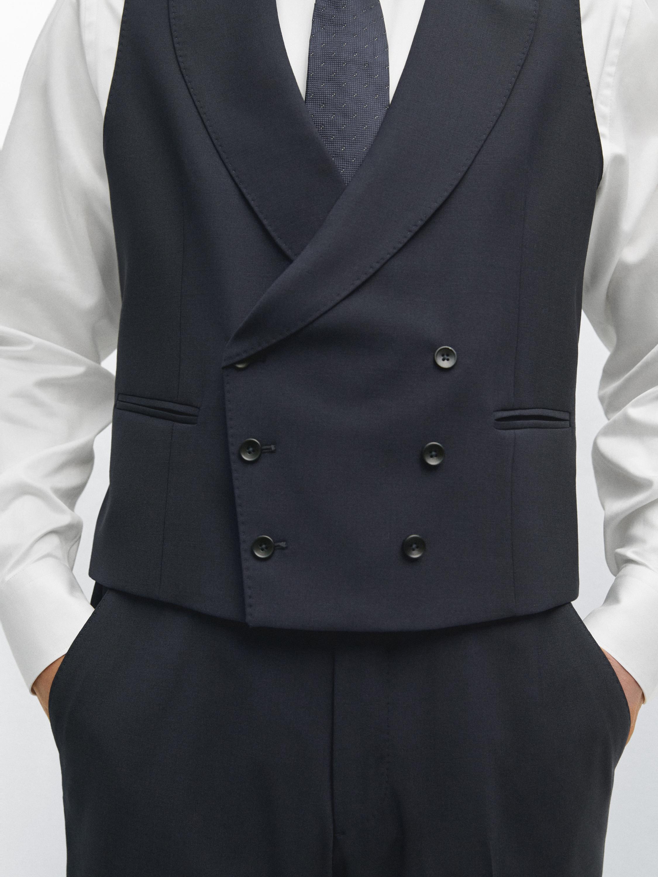 Μεσαίο πλάνο ενός ναυτικού μπλε σταυρωτού γιλέκου με πλαϊνές τσέπες, φορεμένο πάνω από ένα λευκό πουκάμισο και μια μπλε γραβάτα.