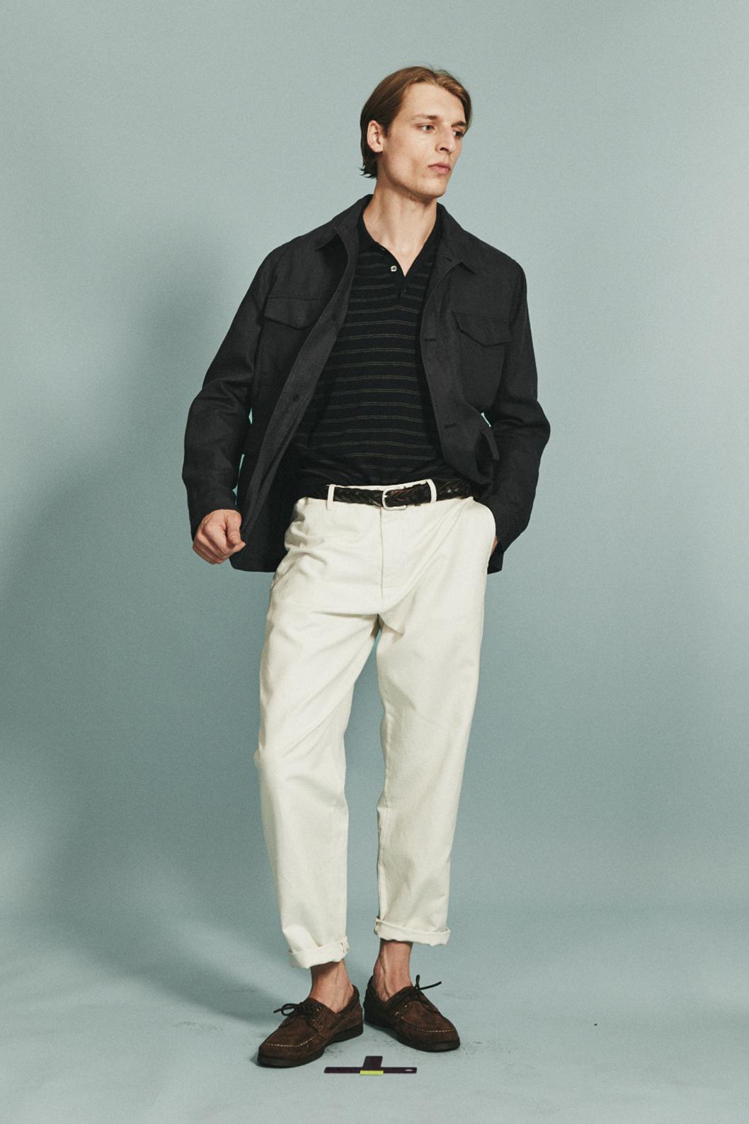 Modèle masculin avec veste noire, t-shirt rayé, pantalon blanc et mocassins marrons. Fond gris clair.