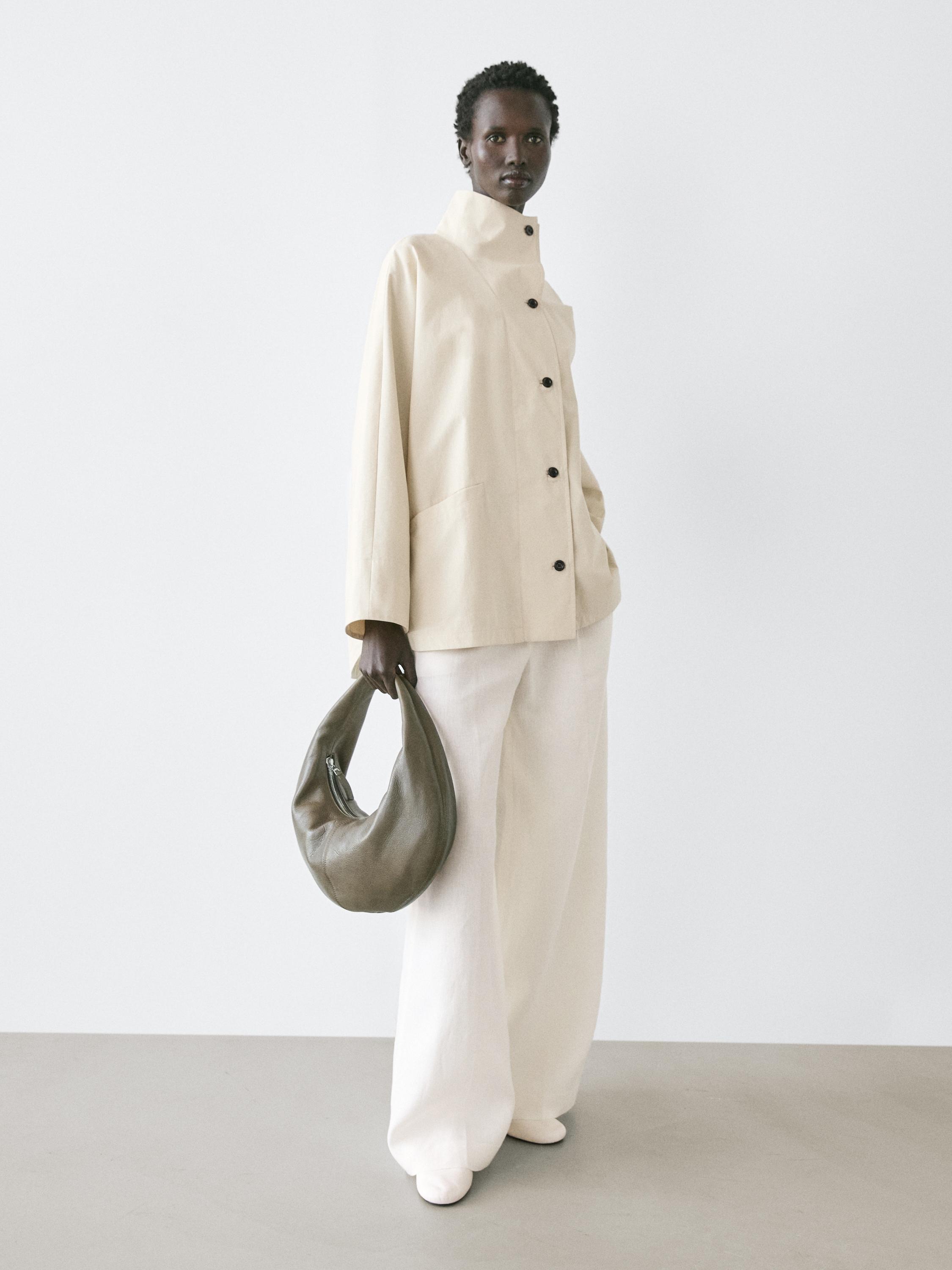 Set van ecru jasje met opstaande kraag en zwarte knopen, ecru wijde broek en bruine tas.