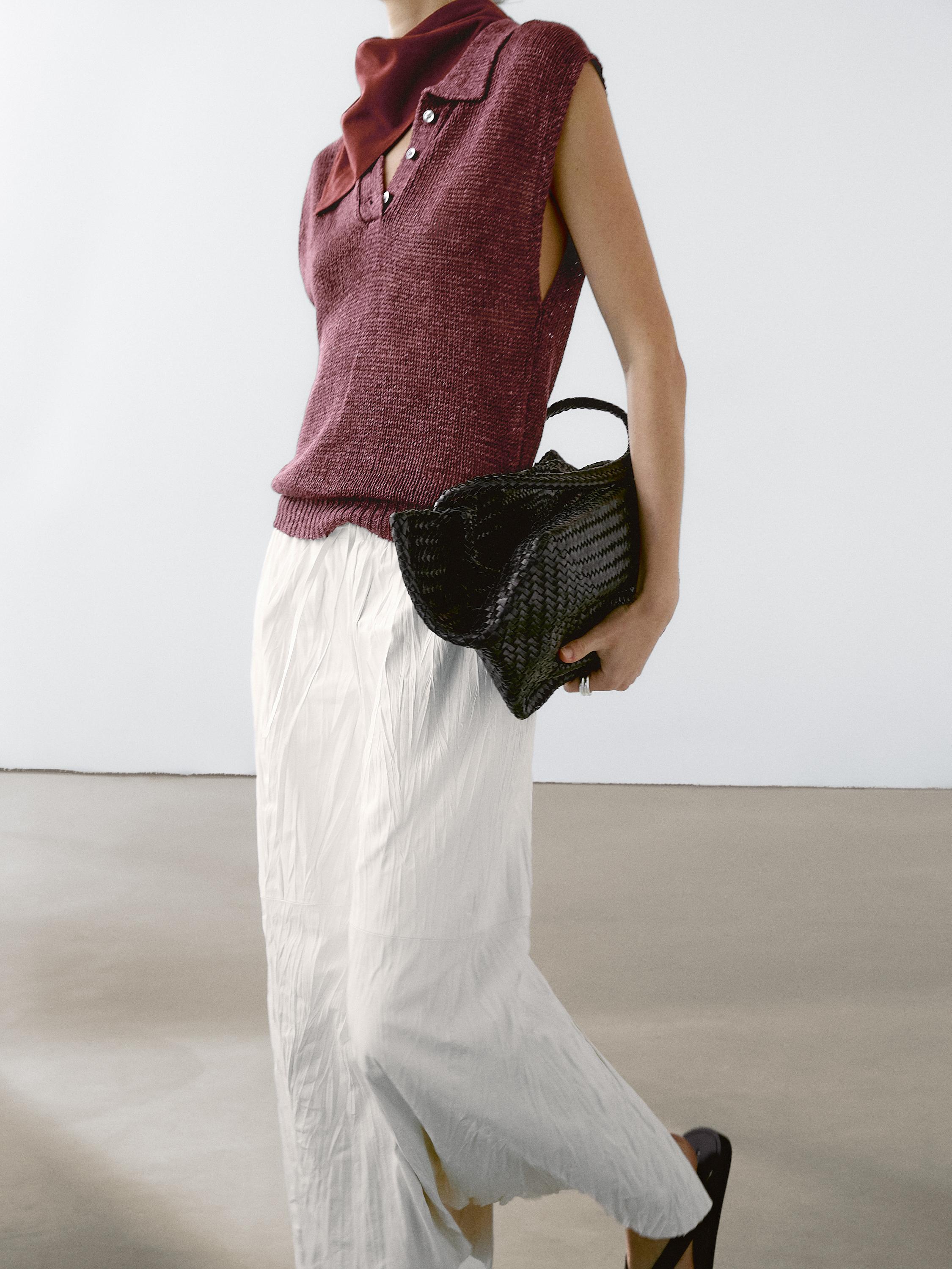 Ensemble composé d'un gilet sans manches bordeaux boutonné, d'une longue jupe blanche plissée et d'un sac noir tressé.