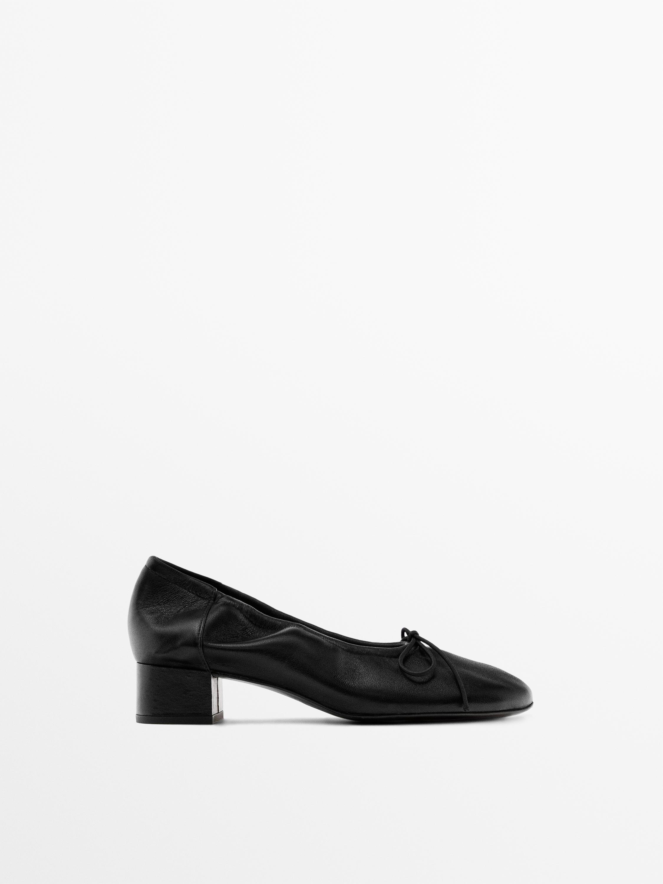 Side view of a black ballet flat with a low heel and a decorative bow on top.