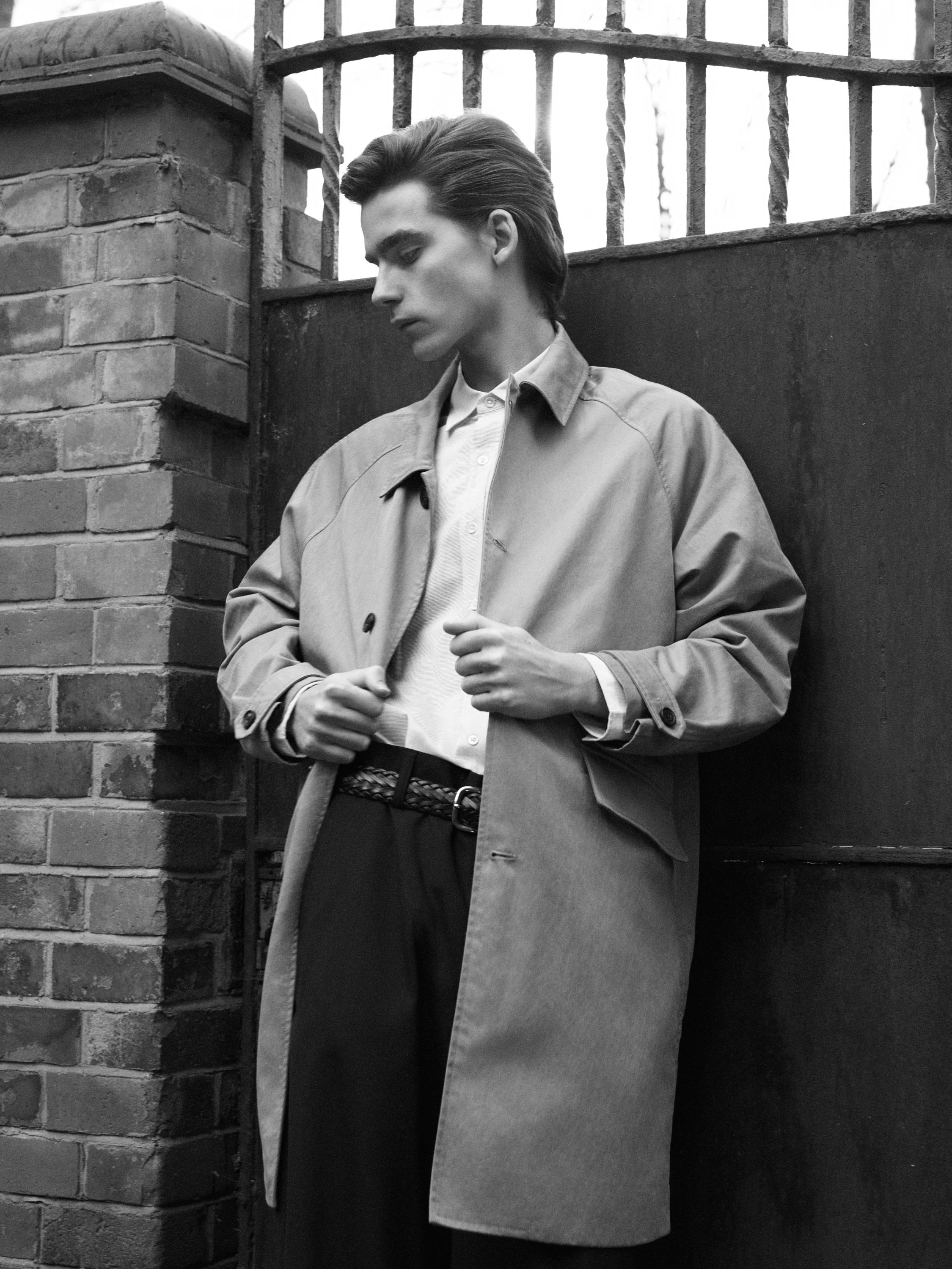 طقم معطف بيج طويل بياقات، وقميص أبيض، وبنطال داكن مع حزام مضفّر.