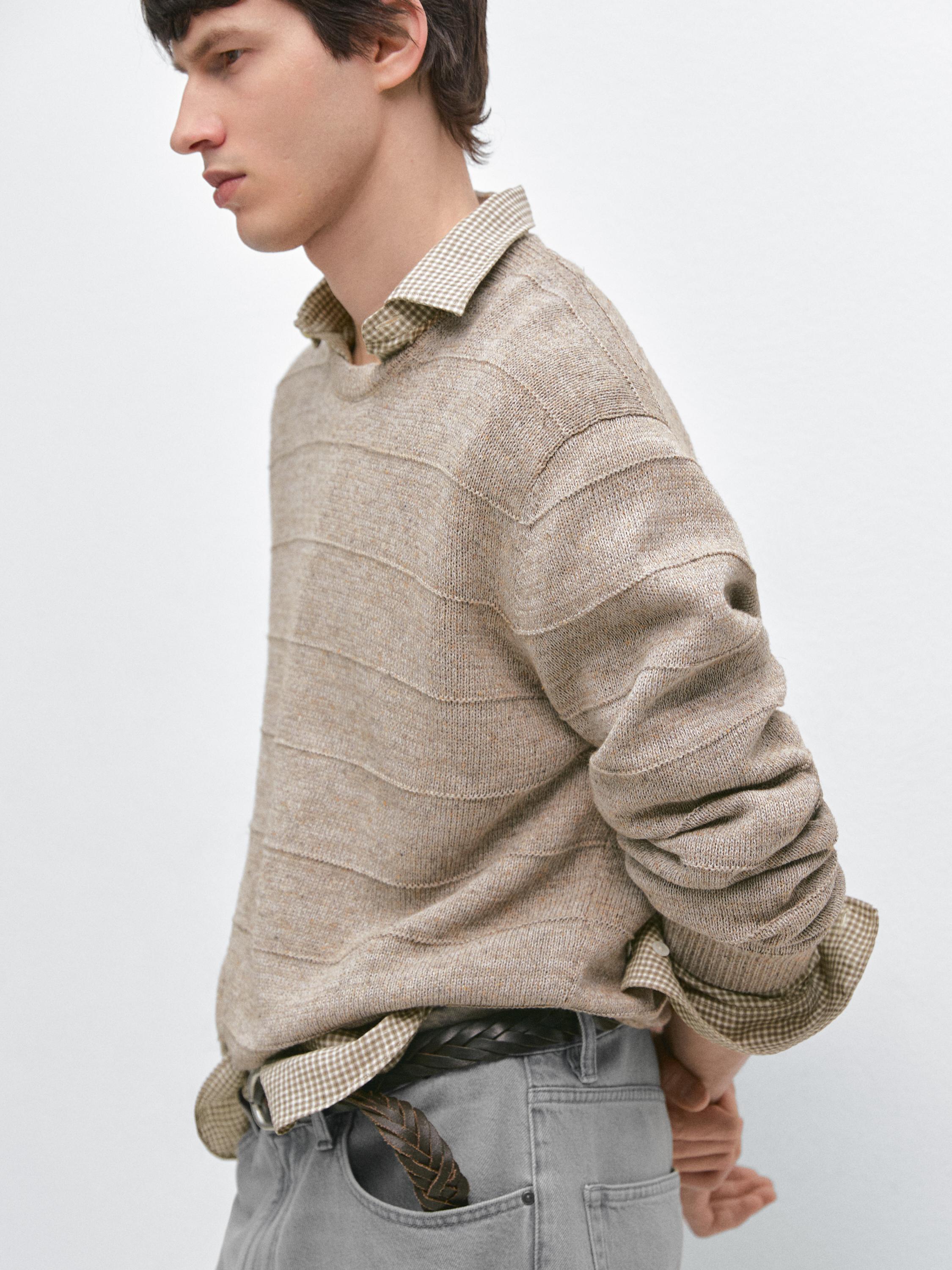 Plano médio de camisola bege com textura de riscas sobre camisa de xadrez castanha. Calças cinzentas com cinto entrançado visível.