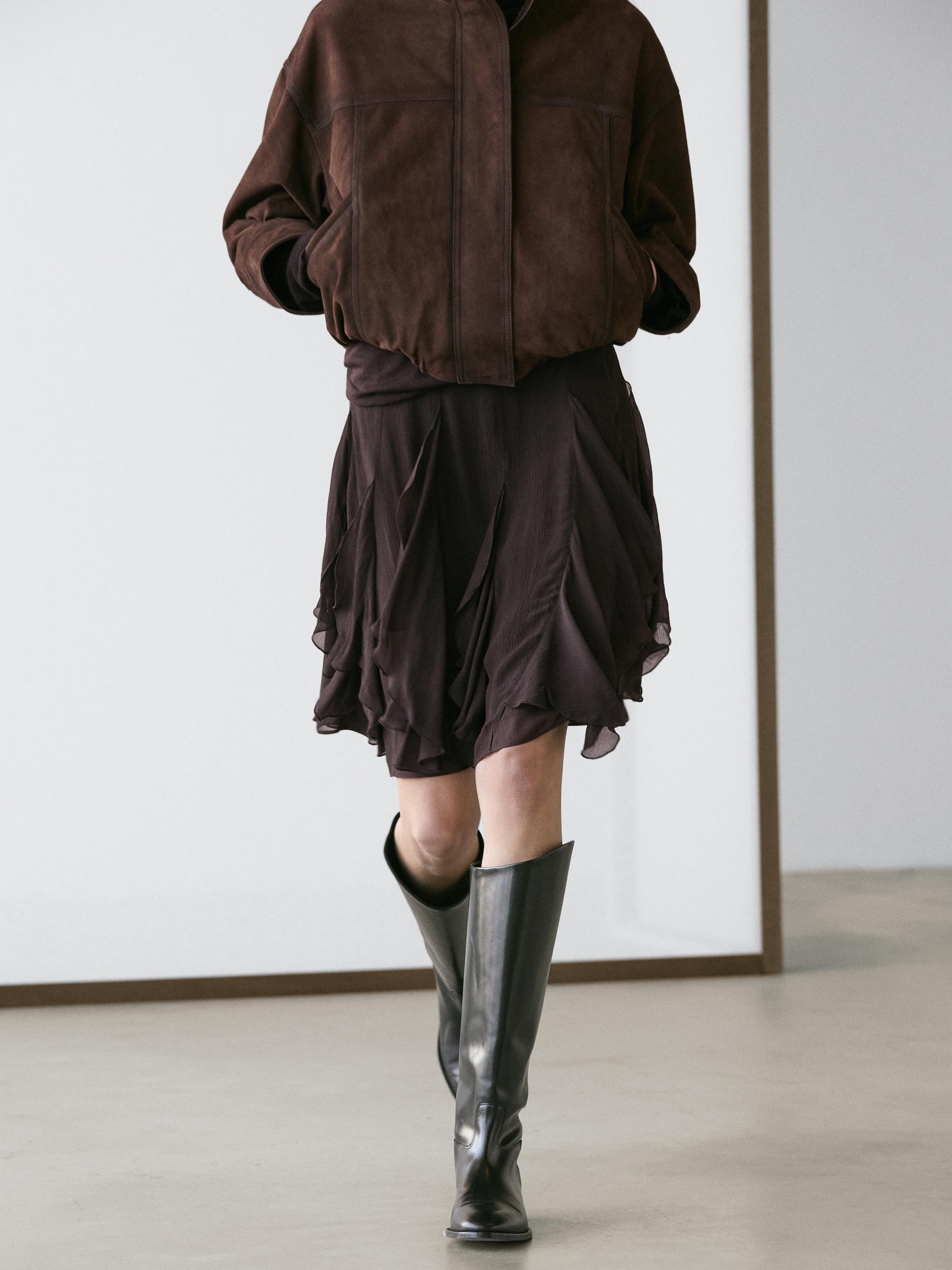 Komplet braon jakne sa džepovima i braon asimetrične suknje sa karnerima, crne duboke čizme.