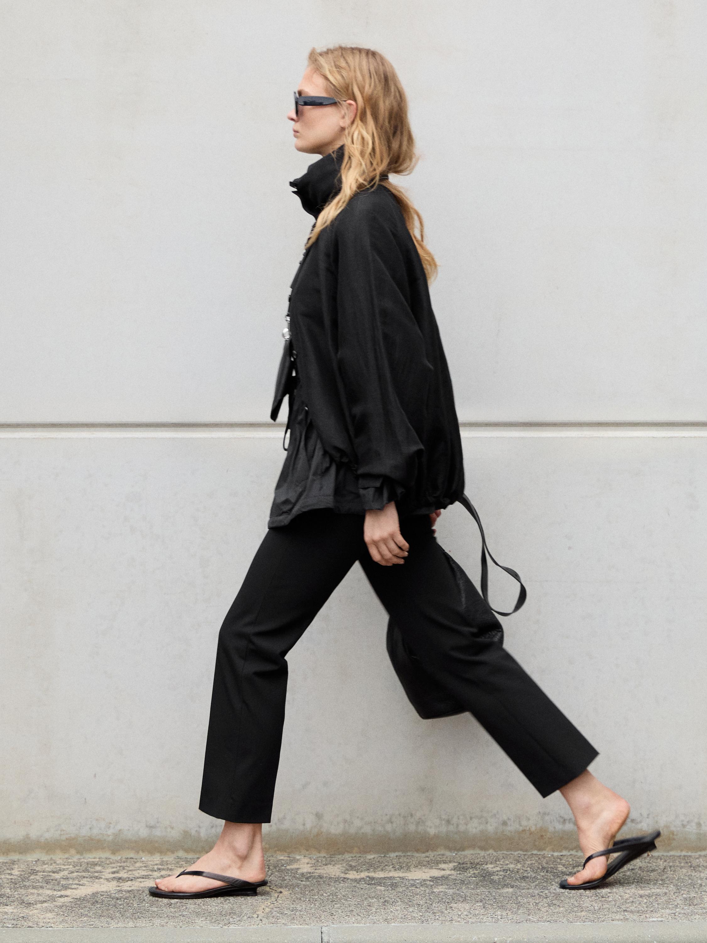 Yüksek yakalı ve geniş kollu siyah ceket takımı, düz siyah pantolon ve siyah sandaletler.