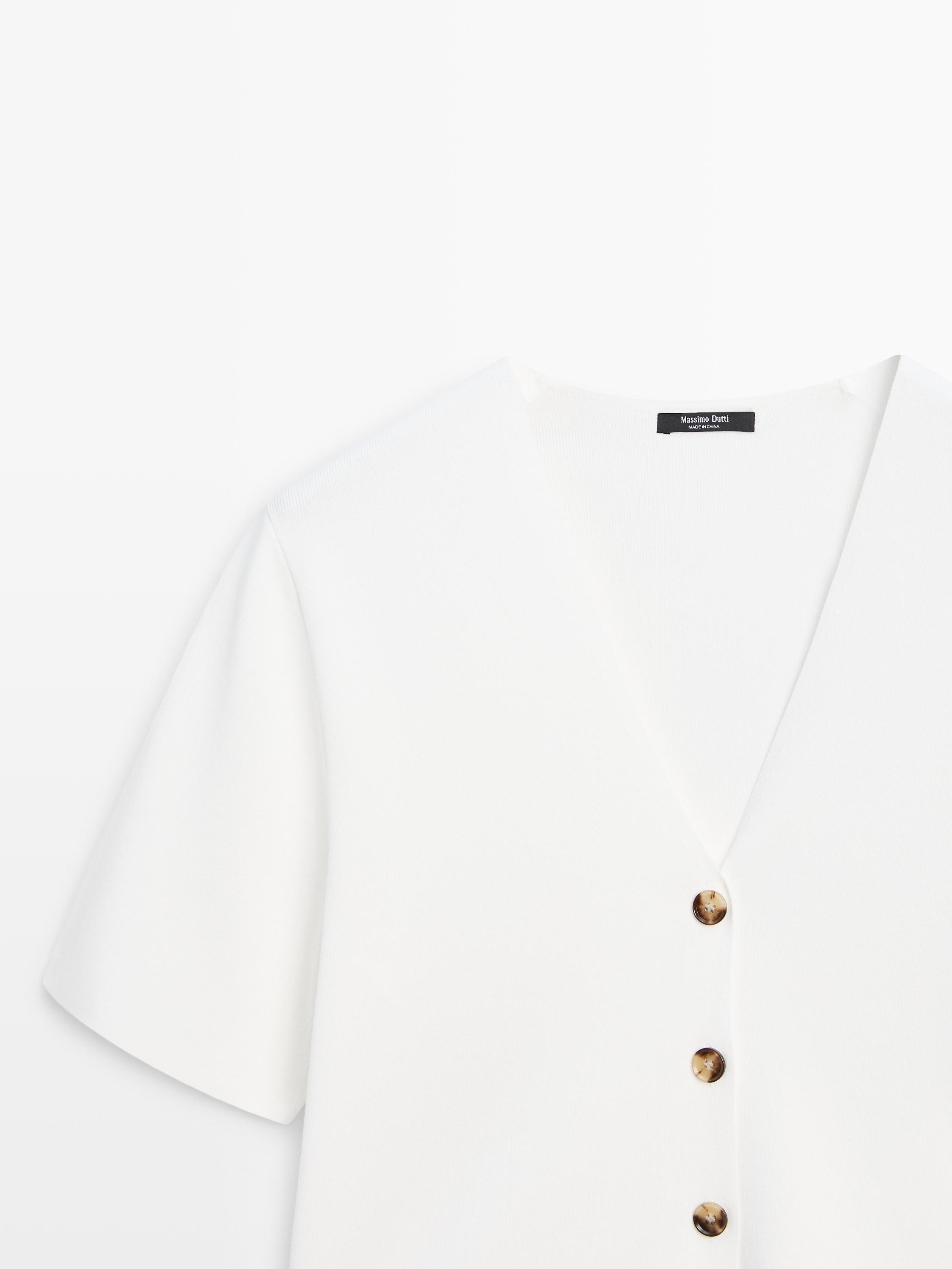 "Detalle de la botonadura de un cárdigan blanco con cuello en V y mangas cortas. Botones marrones visibles."