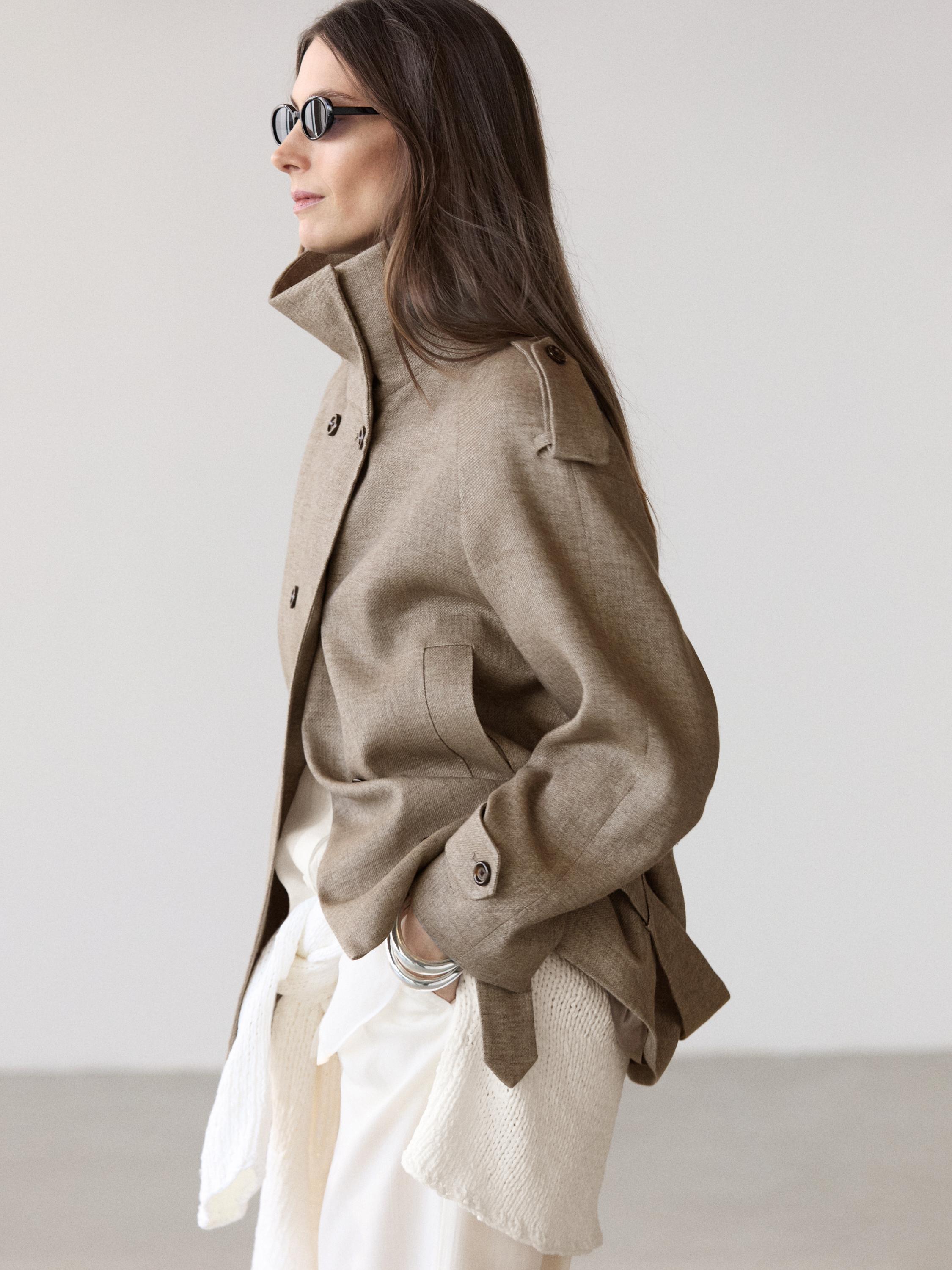 Halflang shot van een taupe grijze blazer met een hoge kraag, knopen aan de voorkant en zijzakken.