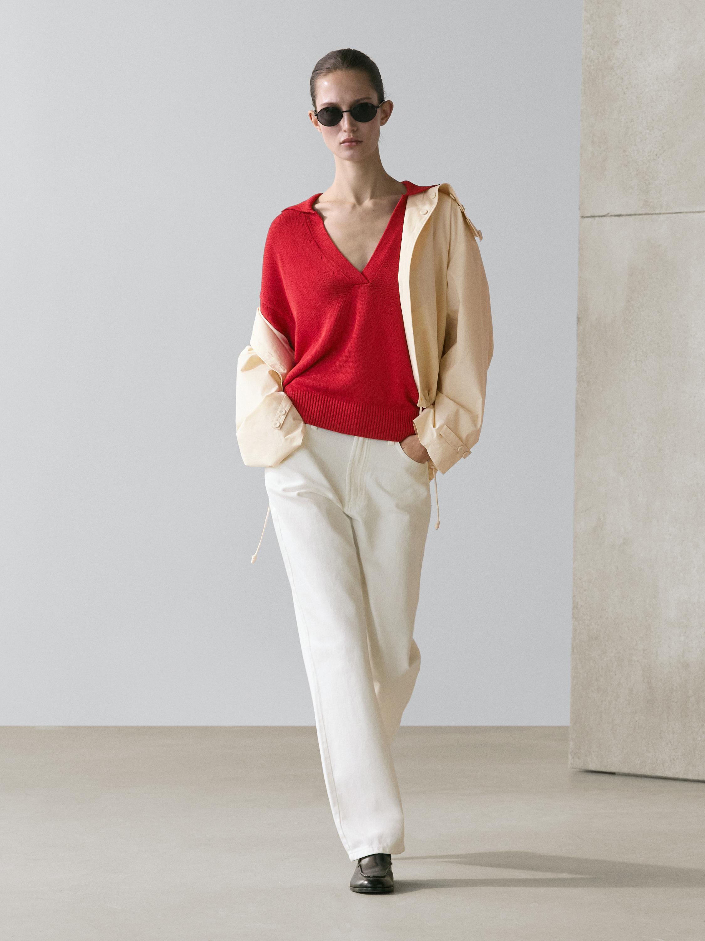 Ensemble composé d'un pull rouge en maille à coupe asymétrique, d'un pantalon blanc droit et d'une veste beige ouverte.