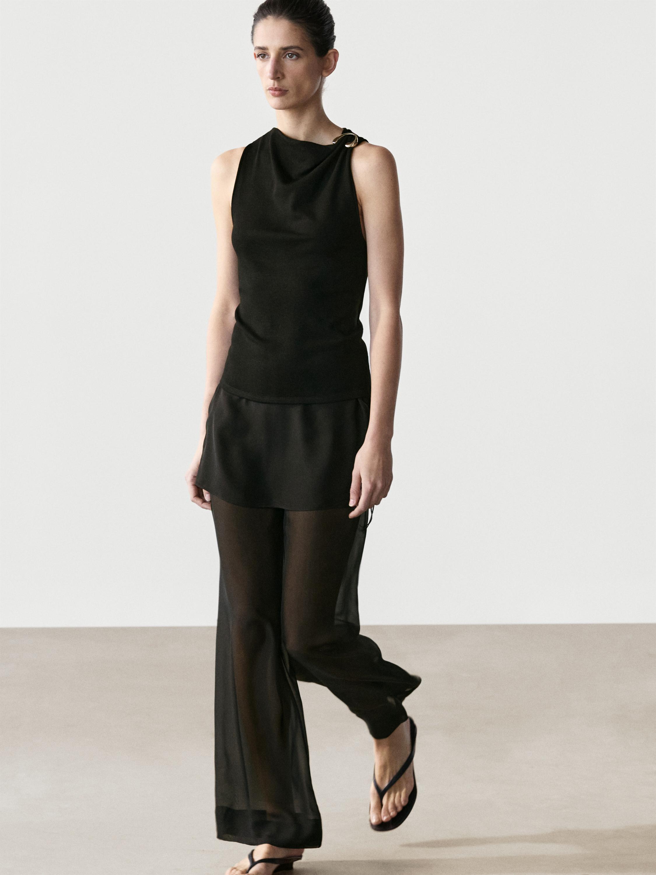 Conjunto de top preto sem mangas com decote drapeado e broche no ombro, calças pretas transparentes e sandálias.