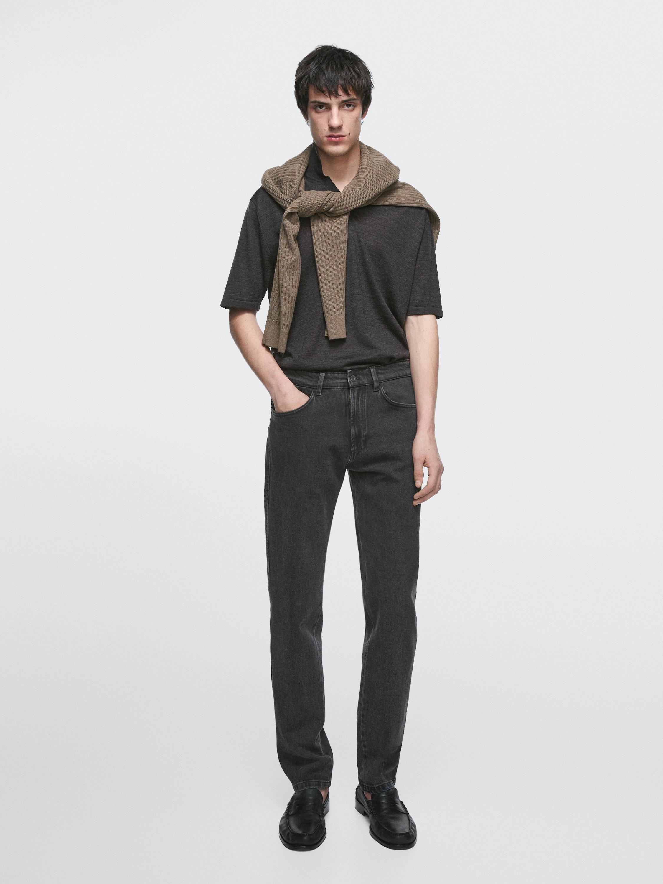 Zestaw składający się z czarnej koszulki, ciemnych jeansów i czarnych butów. Brązowy sweter zawiązany na ramionach.