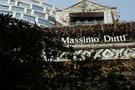 Façade d'un bâtiment ancien avec végétation et panneau "Massimo Dutti". À l'arrière-plan, bâtiment moderne avec fenêtres géométriques.