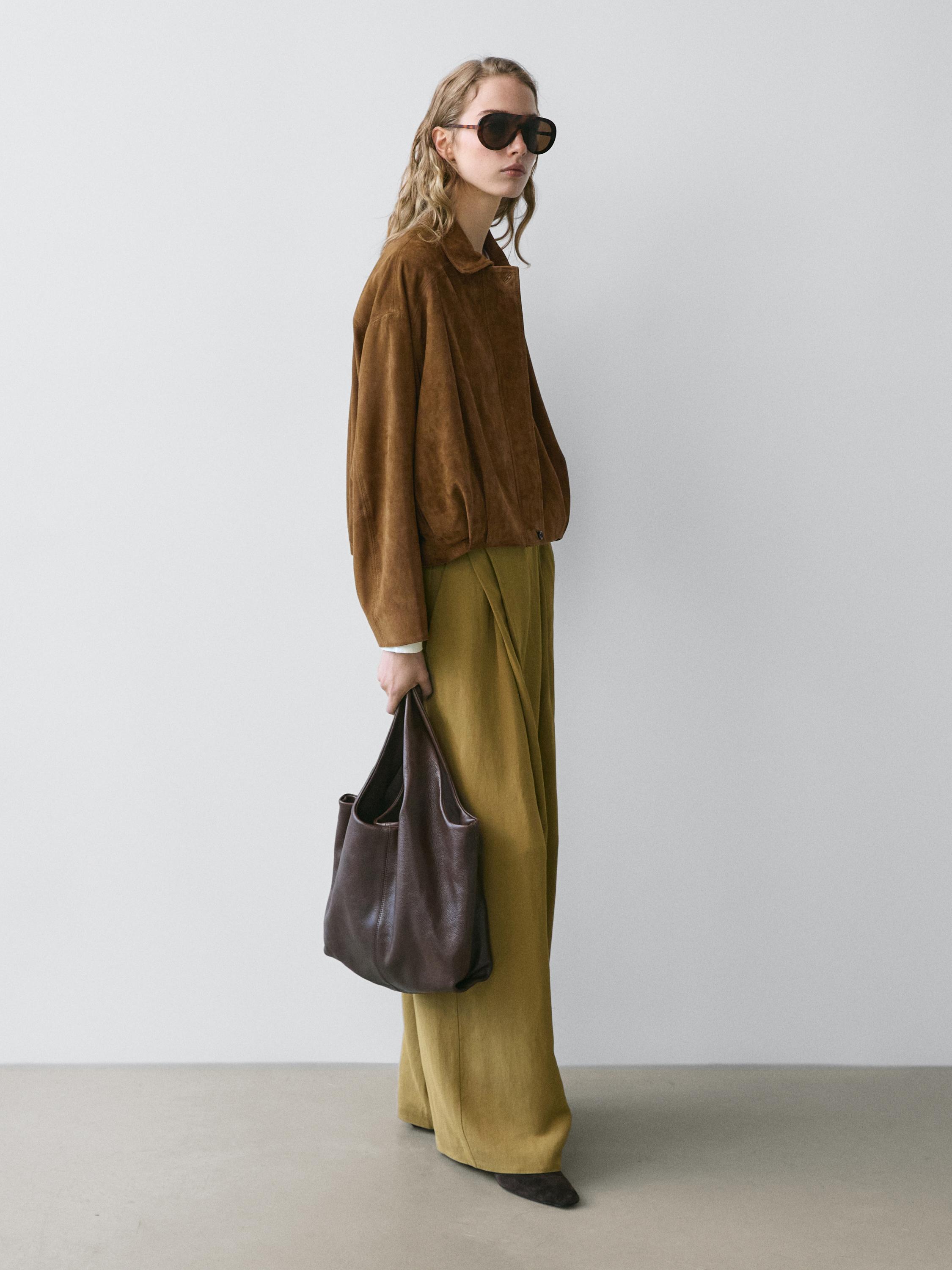 Set aus kurzer brauner Jacke und grünen weiten Hosen. Trägt eine dunkelbraune Tasche und große Sonnenbrillen.